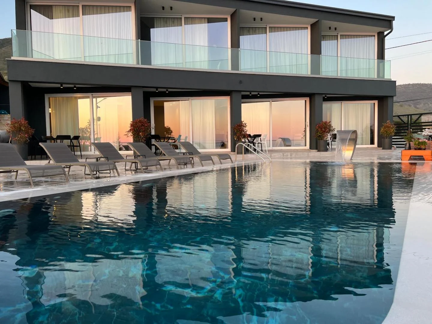 The graphite front of Casa Del Sol at dusk, its lit rooms and a peach sky doubled in the still pool, with grey sun loungers along the coping and a chrome water blade spilling in at the right