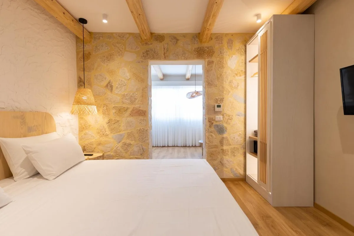 A bed at Dimitri Hotel against a wall of honey-coloured limestone rubble under pine beams, with a fringed cane pendant beside the pillows