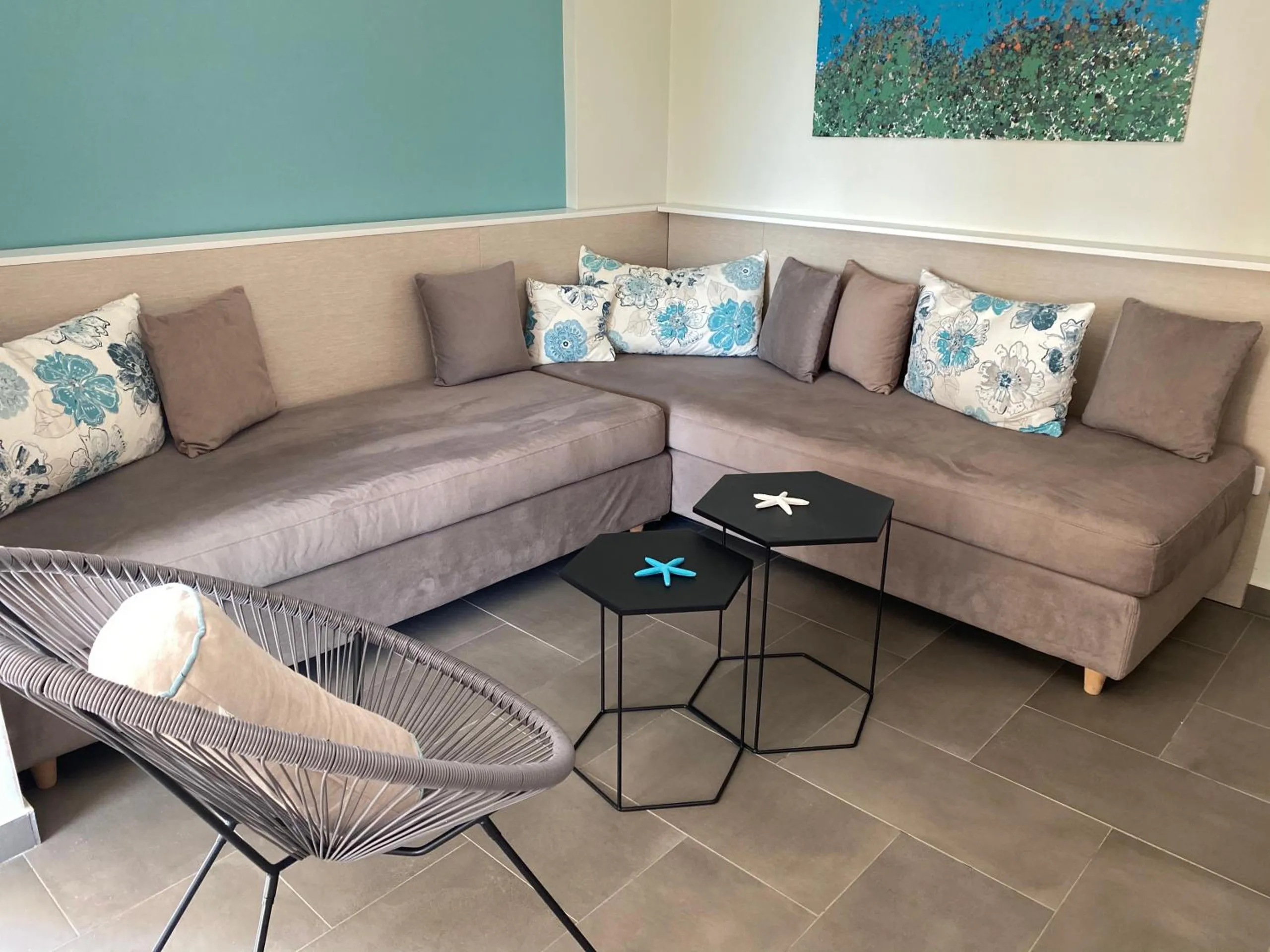 A grey corner sofa with blue flowered cushions, a wire chair and two hexagon tables on a porcelain floor