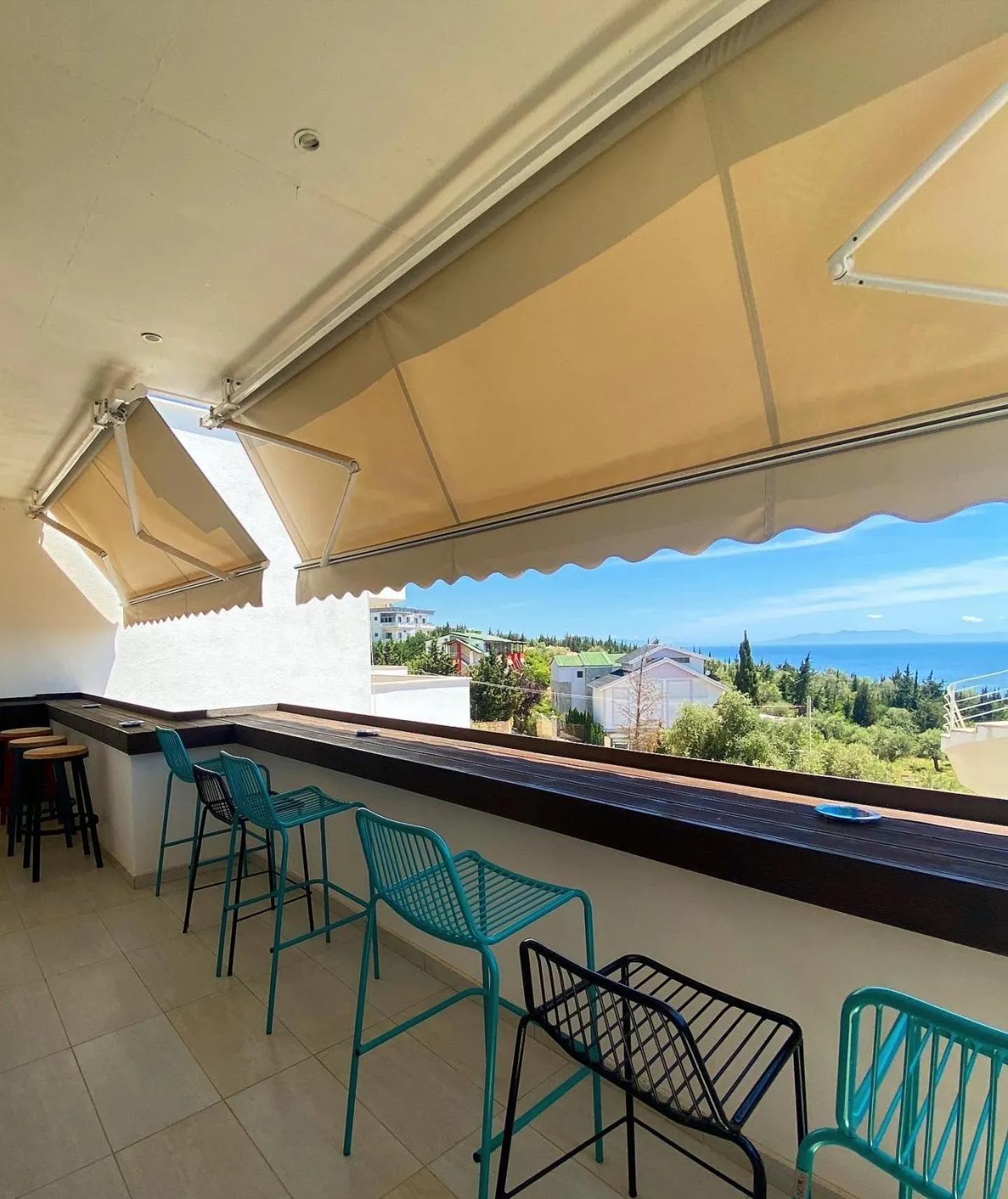 A timber counter with teal bar stools under a striped awning, looking over trees and rooftops to the sea