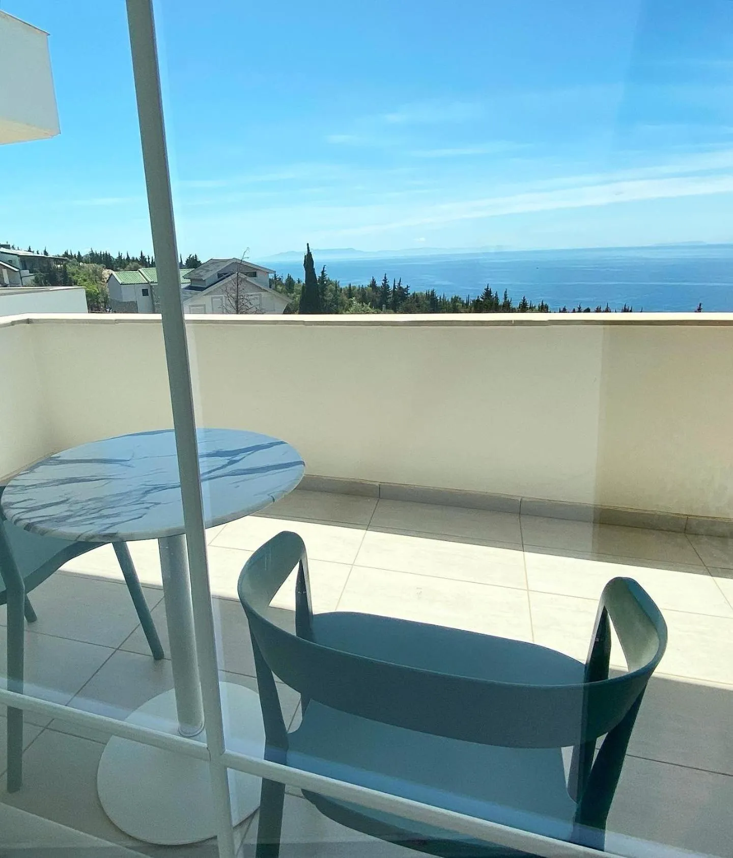 Two petrol-blue chairs at a marble-topped table on a white balcony, the sea beyond the trees