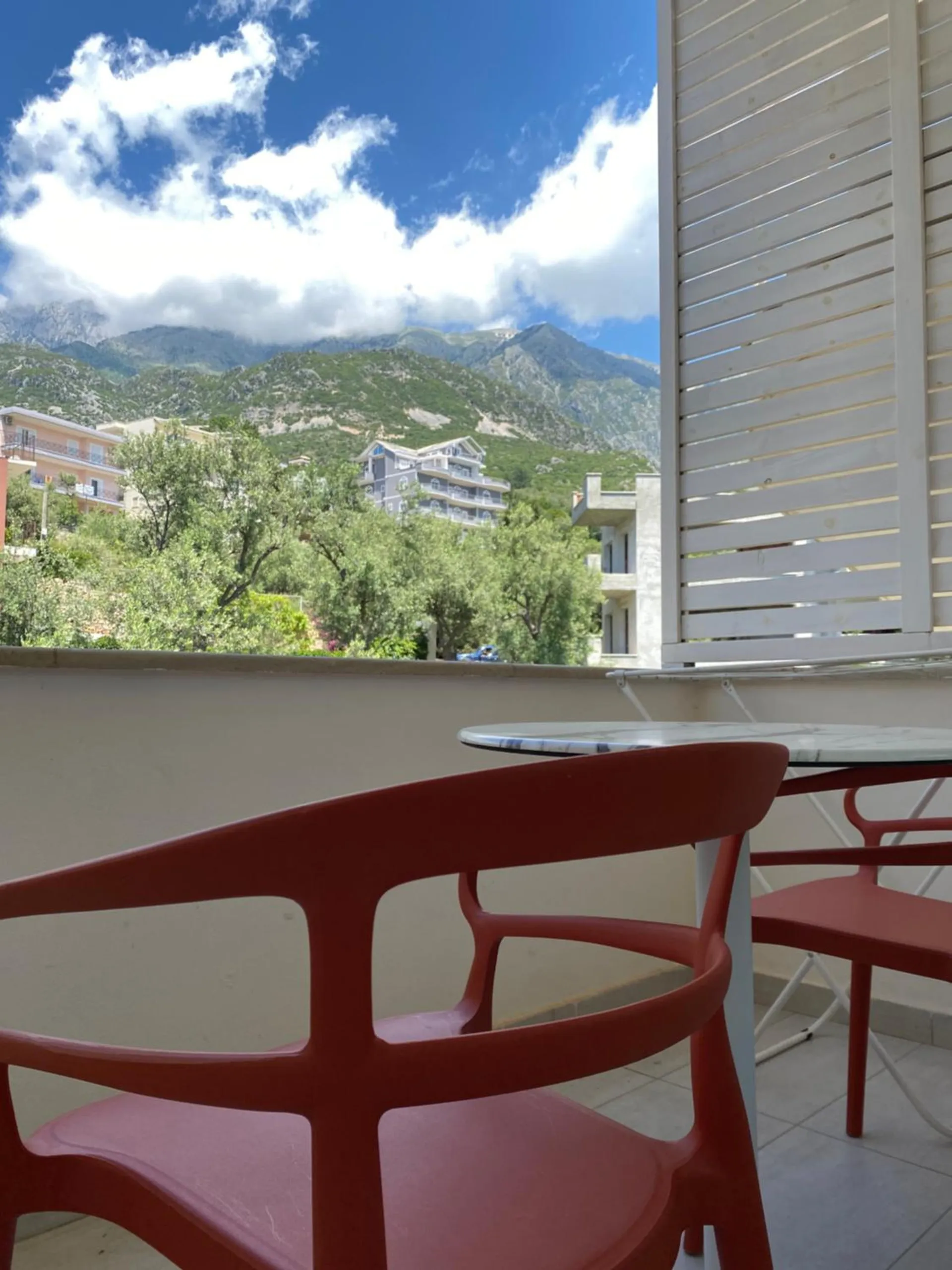 Red chairs at a balcony table, olive trees and white buildings below a green mountainside