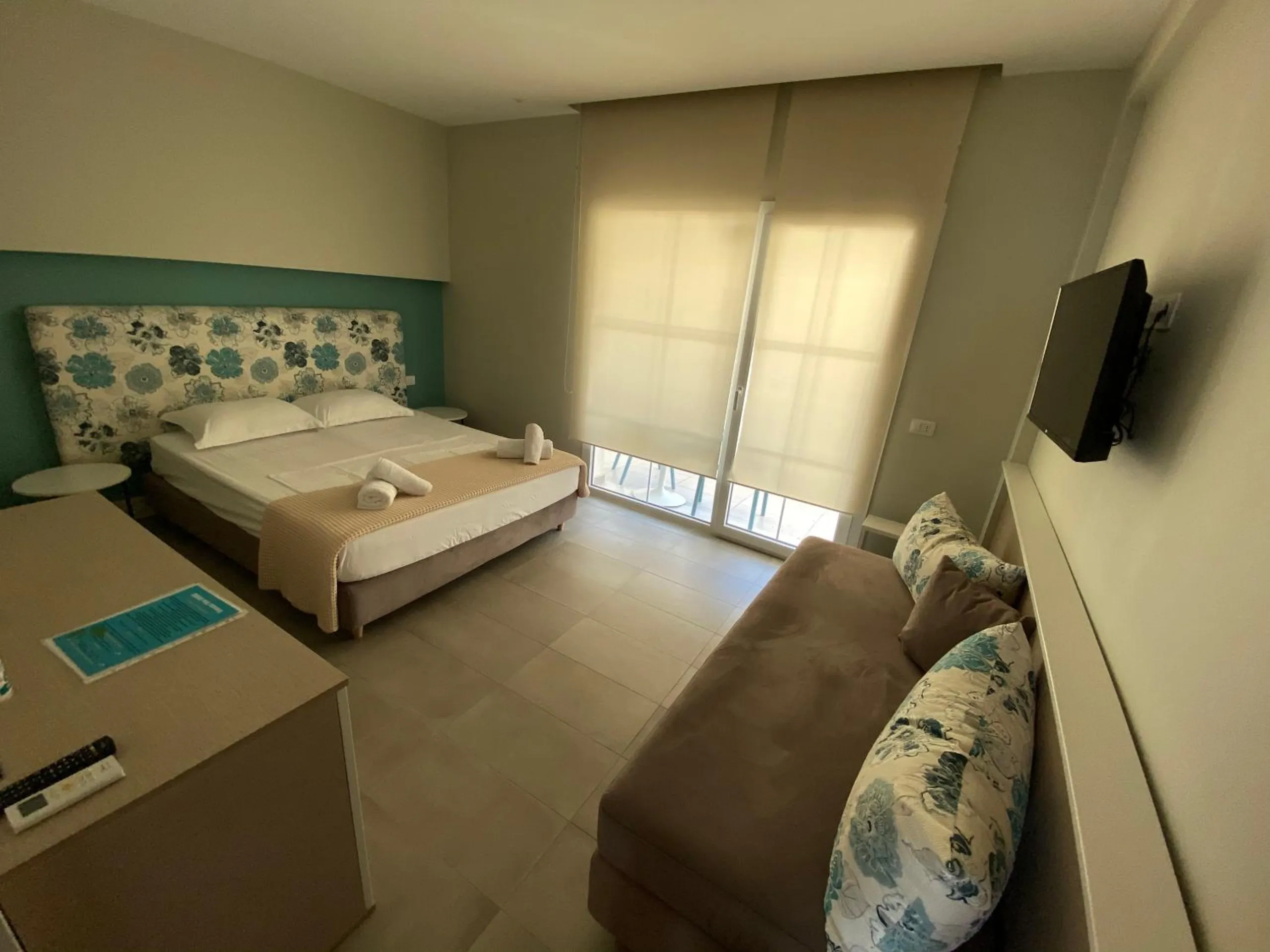 A wide room with a bed on the teal wall, a long sofa with flowered cushions and a roller blind over the balcony doors