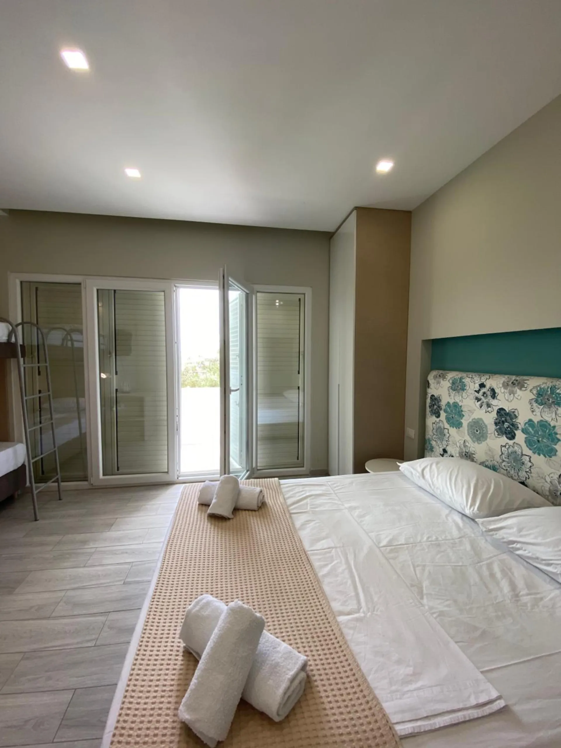 A queen bed under a flowered headboard, bunk beds by the open balcony doors and greenery outside