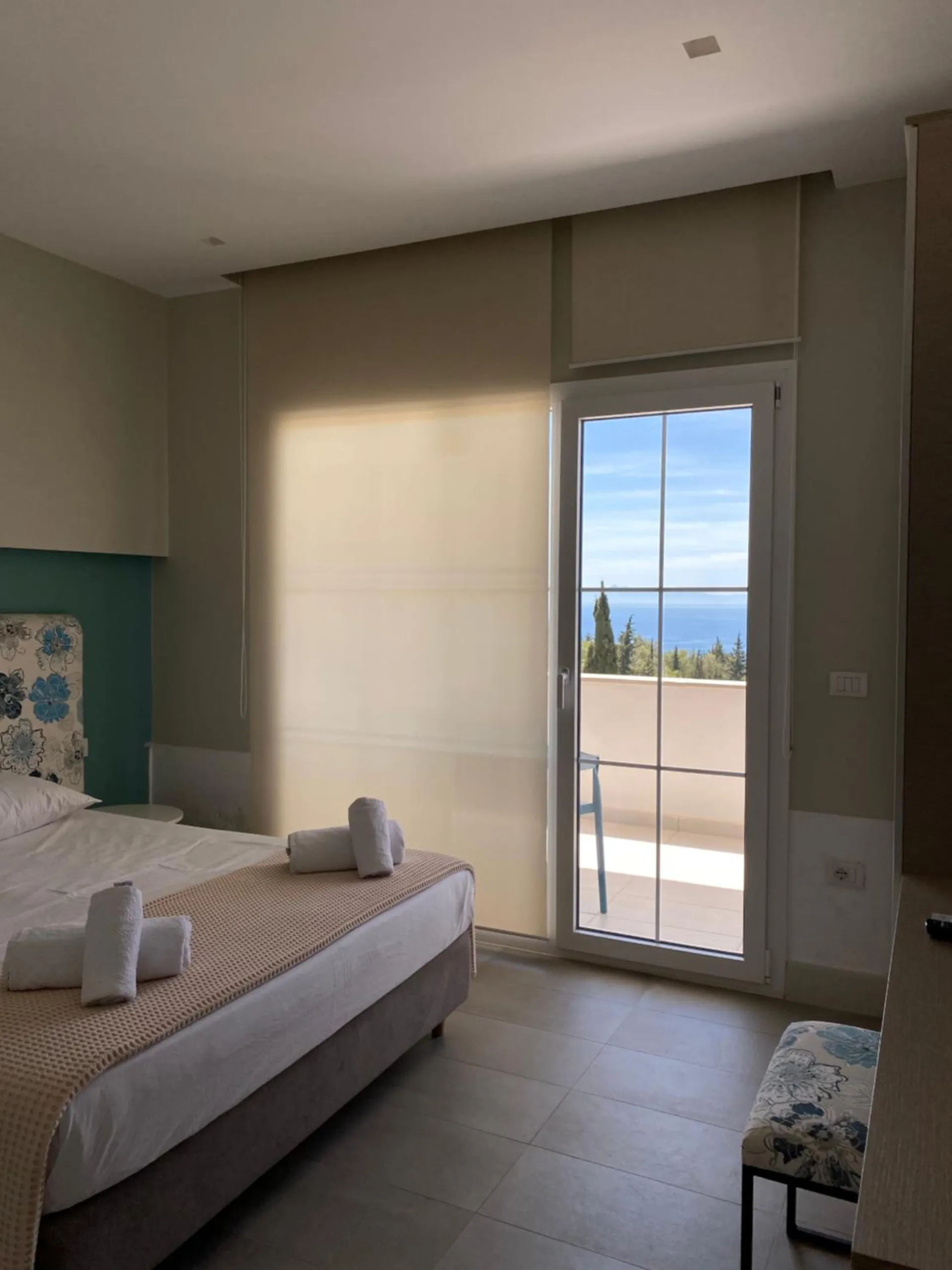 A room with a teal wall, a bed with rolled towels and a glass door onto a balcony with the sea and cypresses beyond