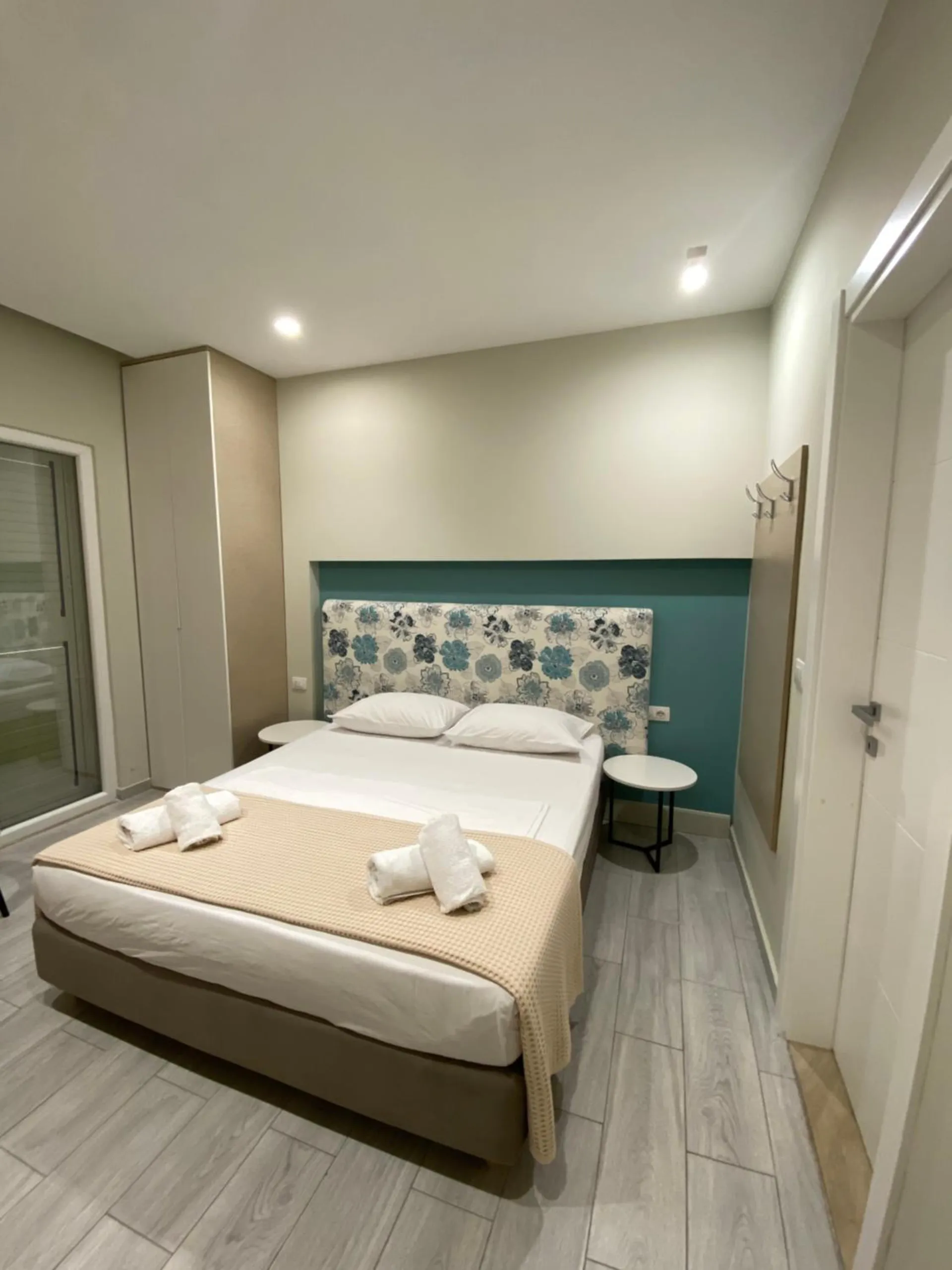 A double bed with rolled towels in front of a teal wall and a flowered headboard, a glass door to one side