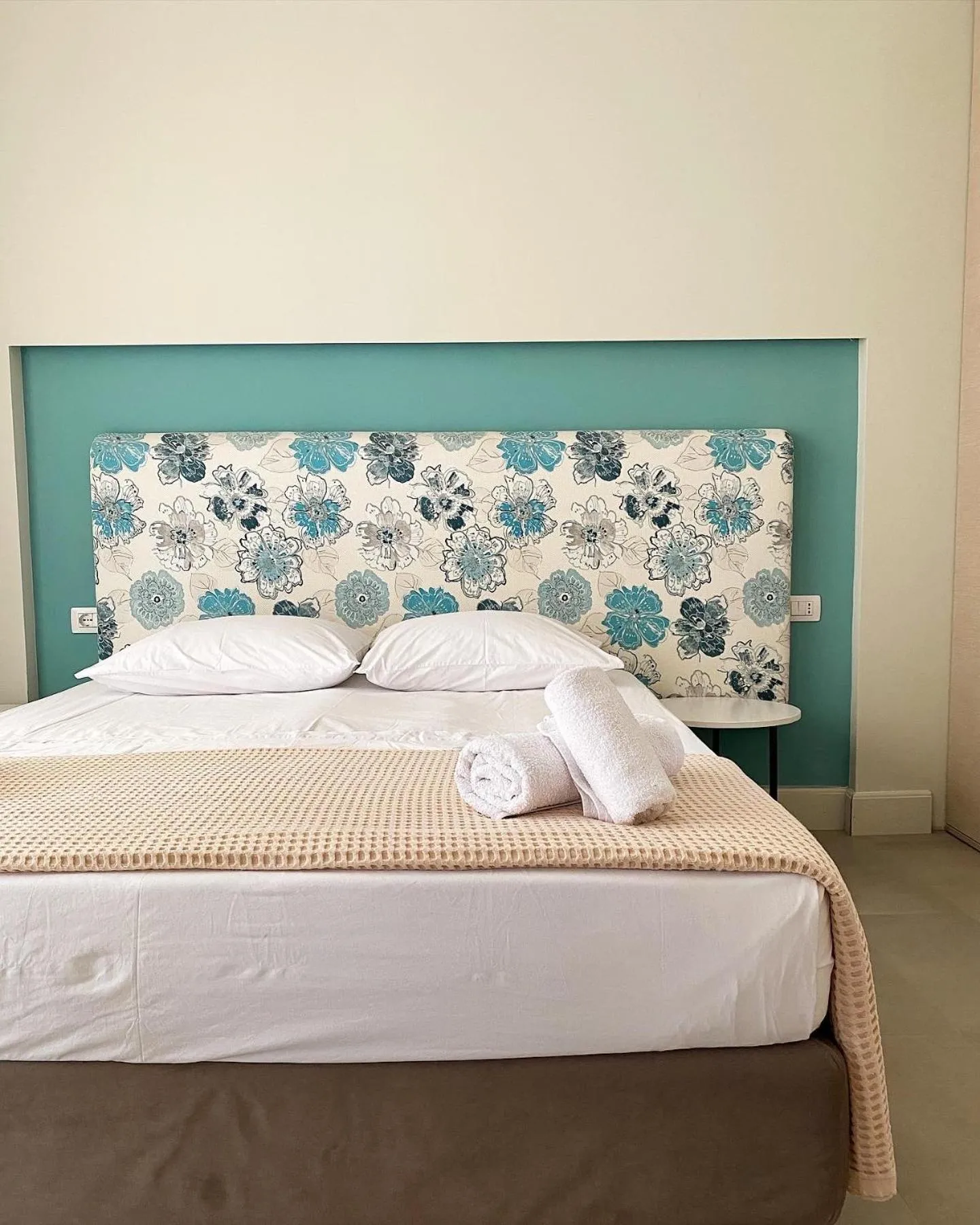 A queen bed with rolled towels in front of a teal wall and a headboard printed with blue flowers