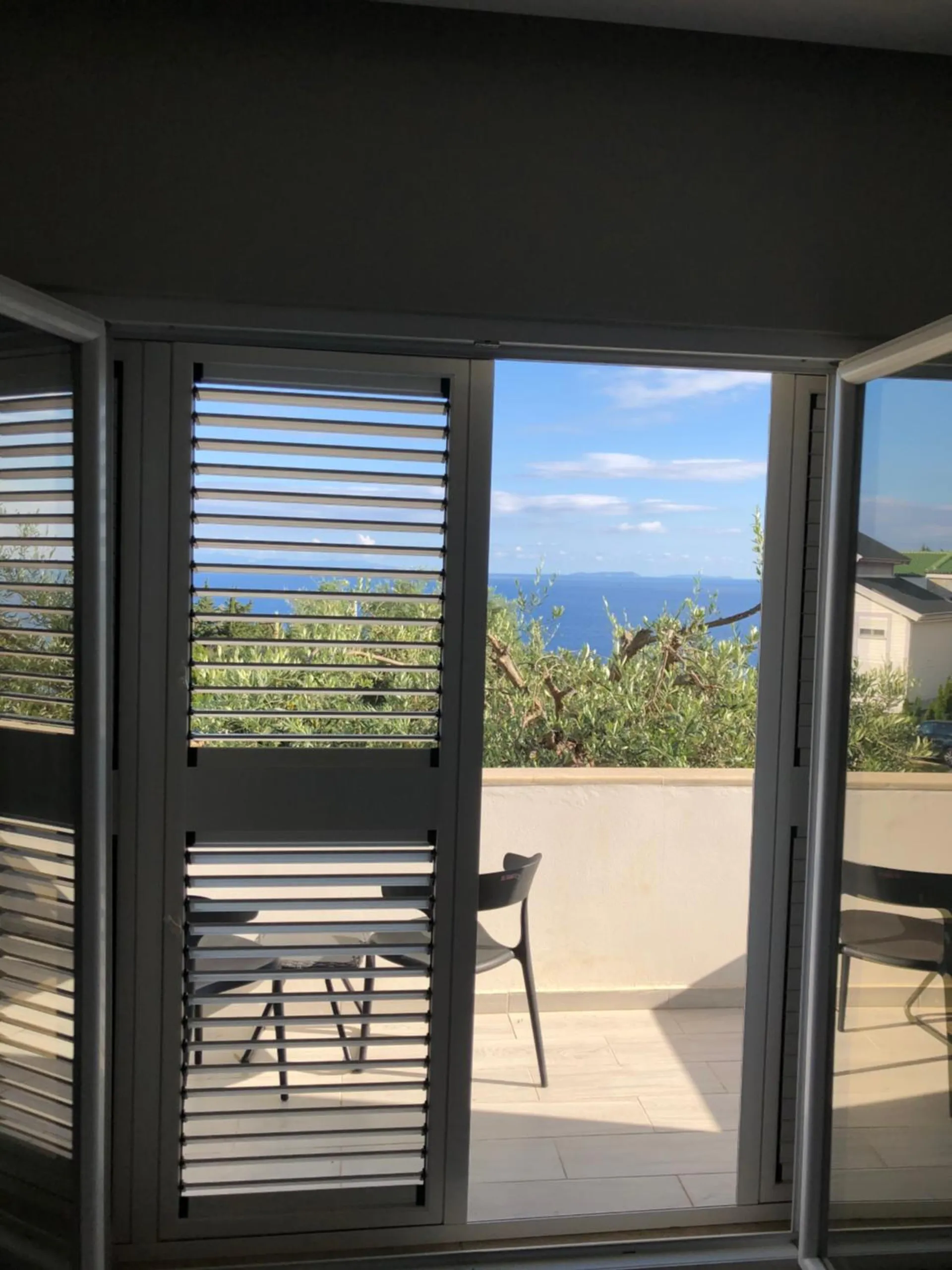Grey louvred doors folded open onto a white terrace wall, olive trees and the sea beyond