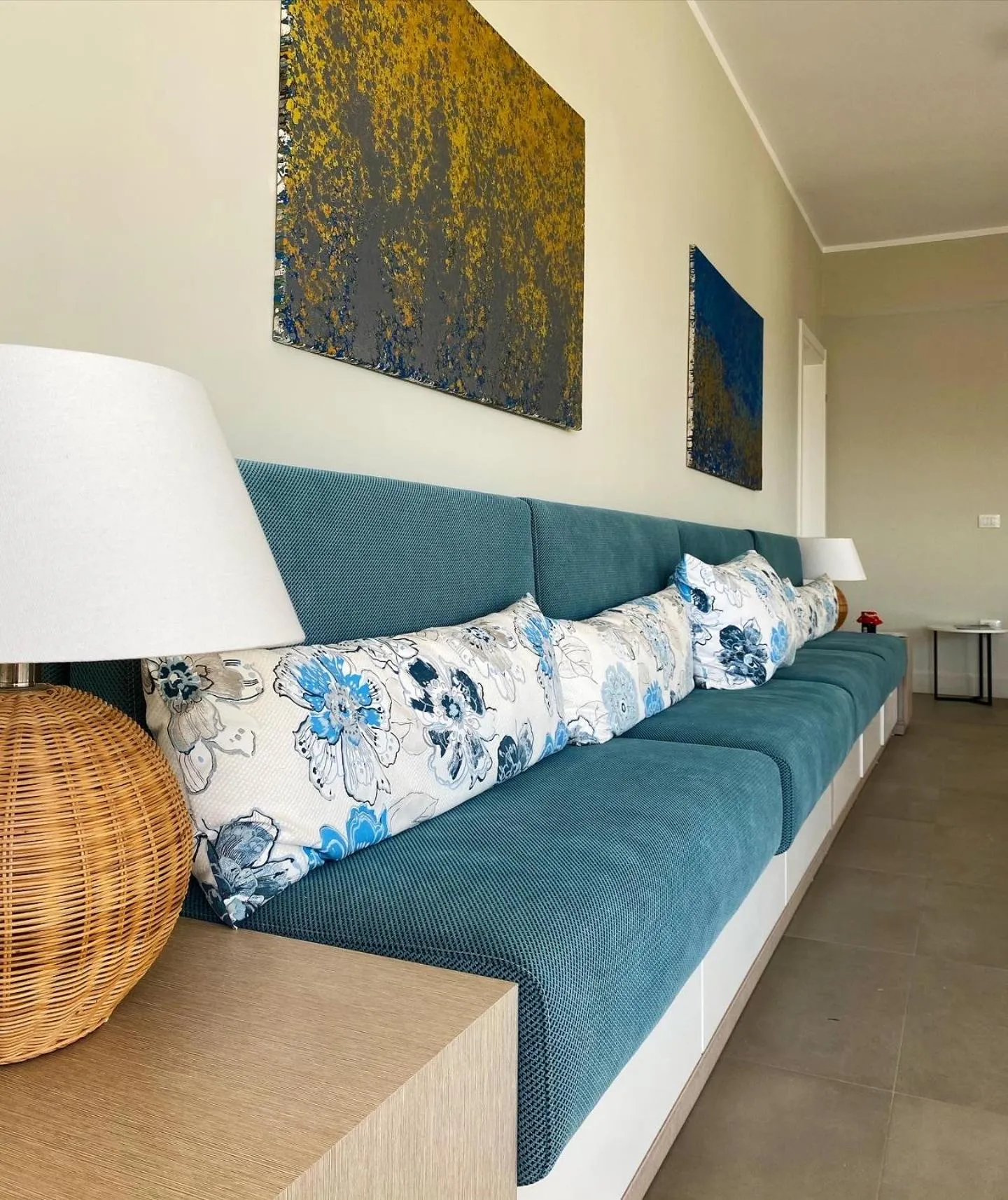 A built-in teal sofa with blue flowered cushions, a wicker lamp and a round side table