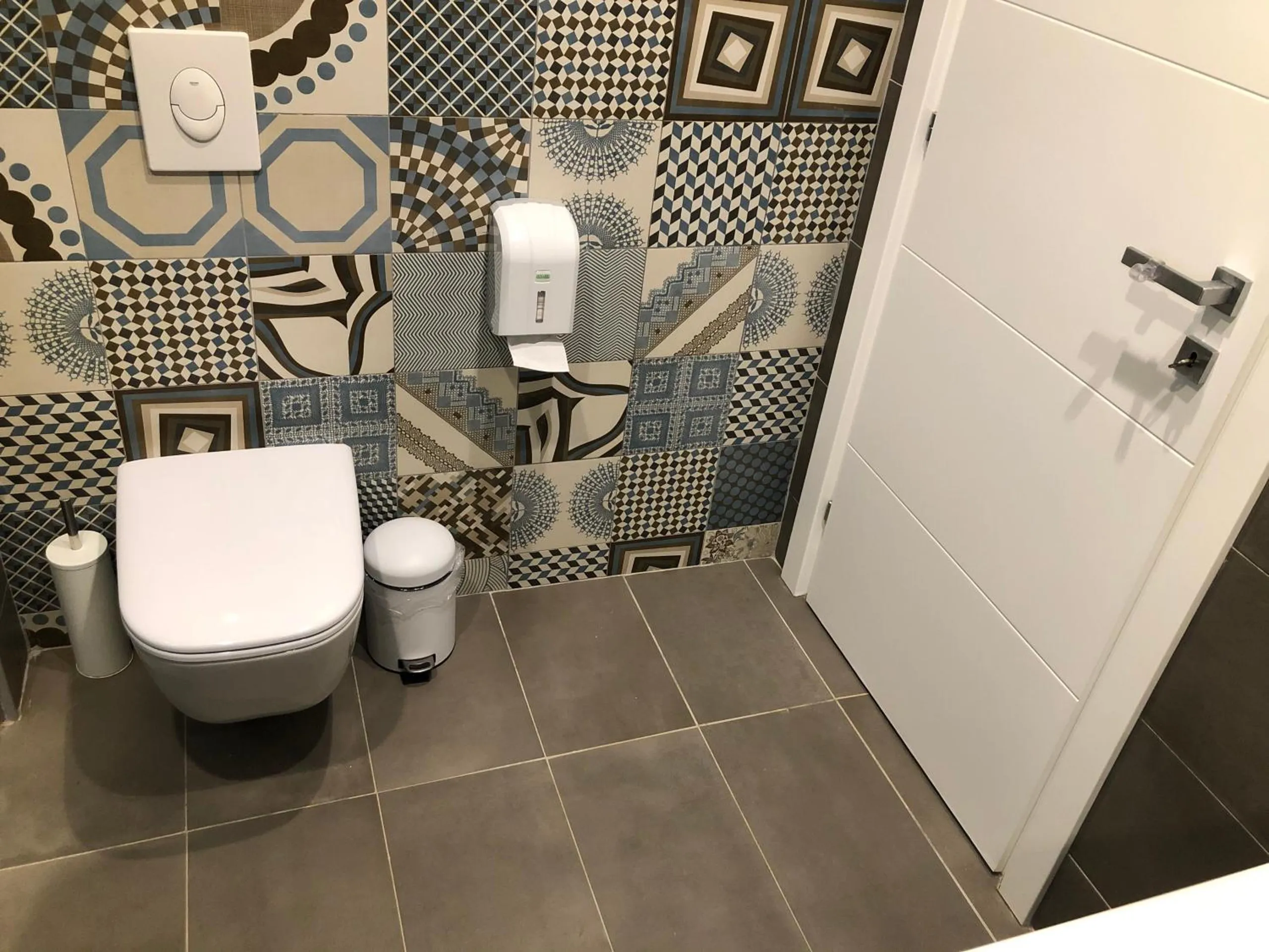 A bathroom with a wall-hung toilet in front of a wall of patterned tiles in blue, grey and sand