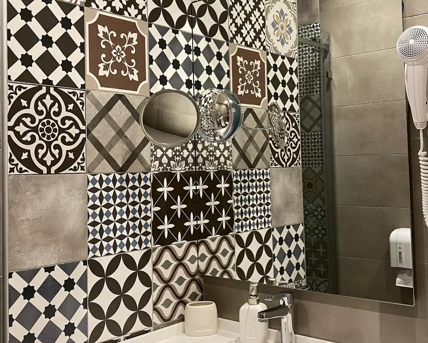 A bathroom wall of patterned tiles in black, white, brown and blue around a mirror and a round shaving mirror