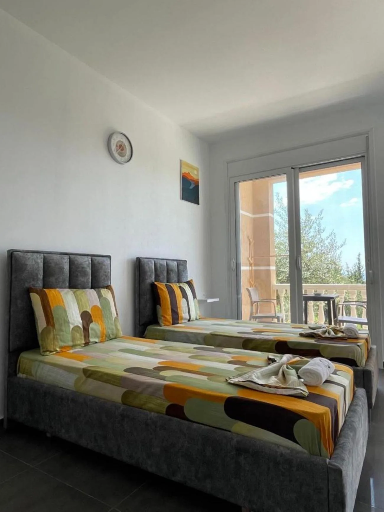 Two single beds with patterned covers beside a glass door onto a porch with a table, chairs and olive trees