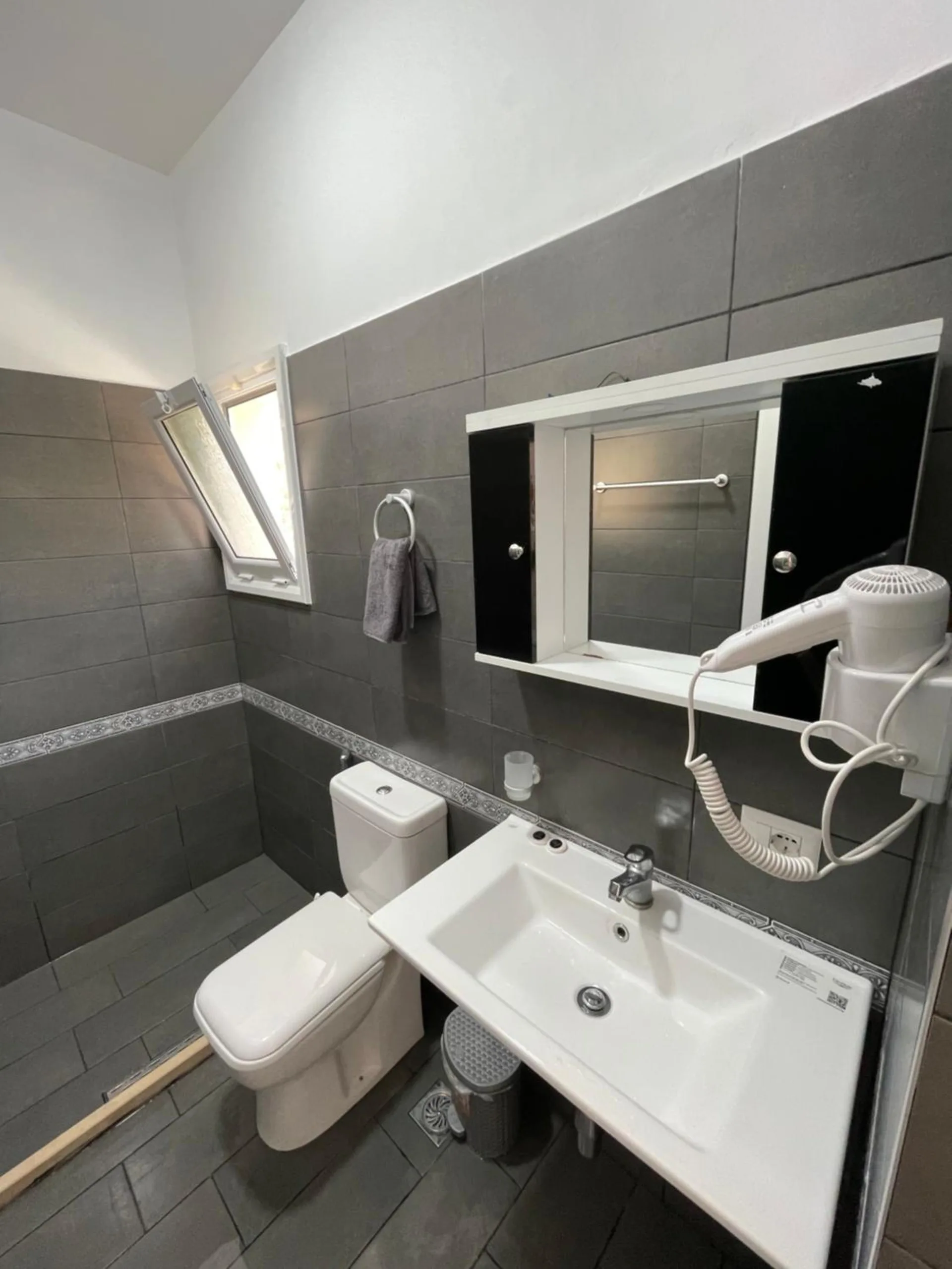 A shower room in dark grey tile with a white basin, a toilet, a mirror cabinet and a hairdryer