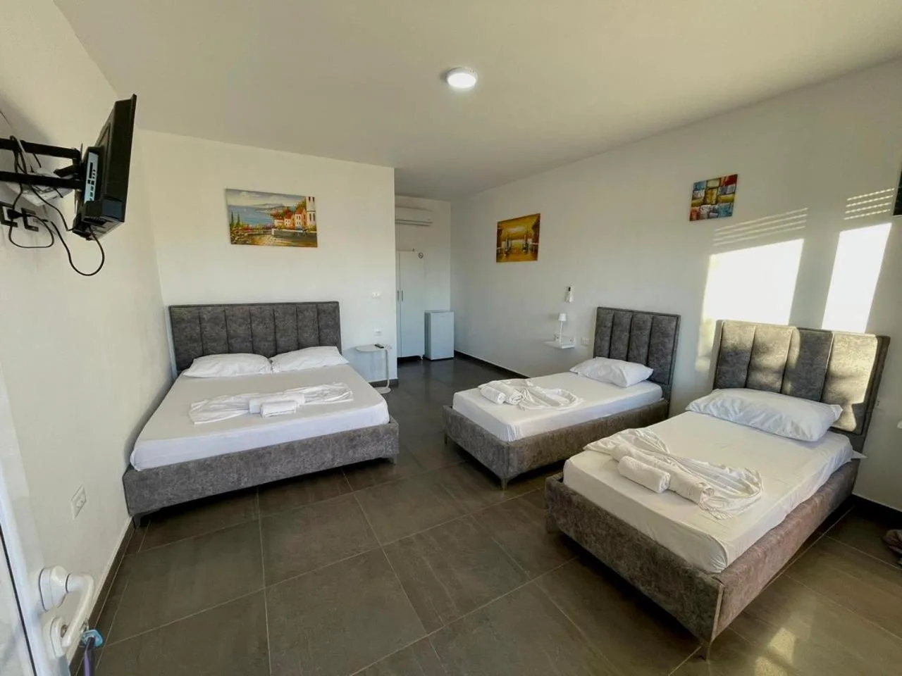 Morning light on a double bed and two single beds in grey frames on a dark tiled floor, with paintings on white walls