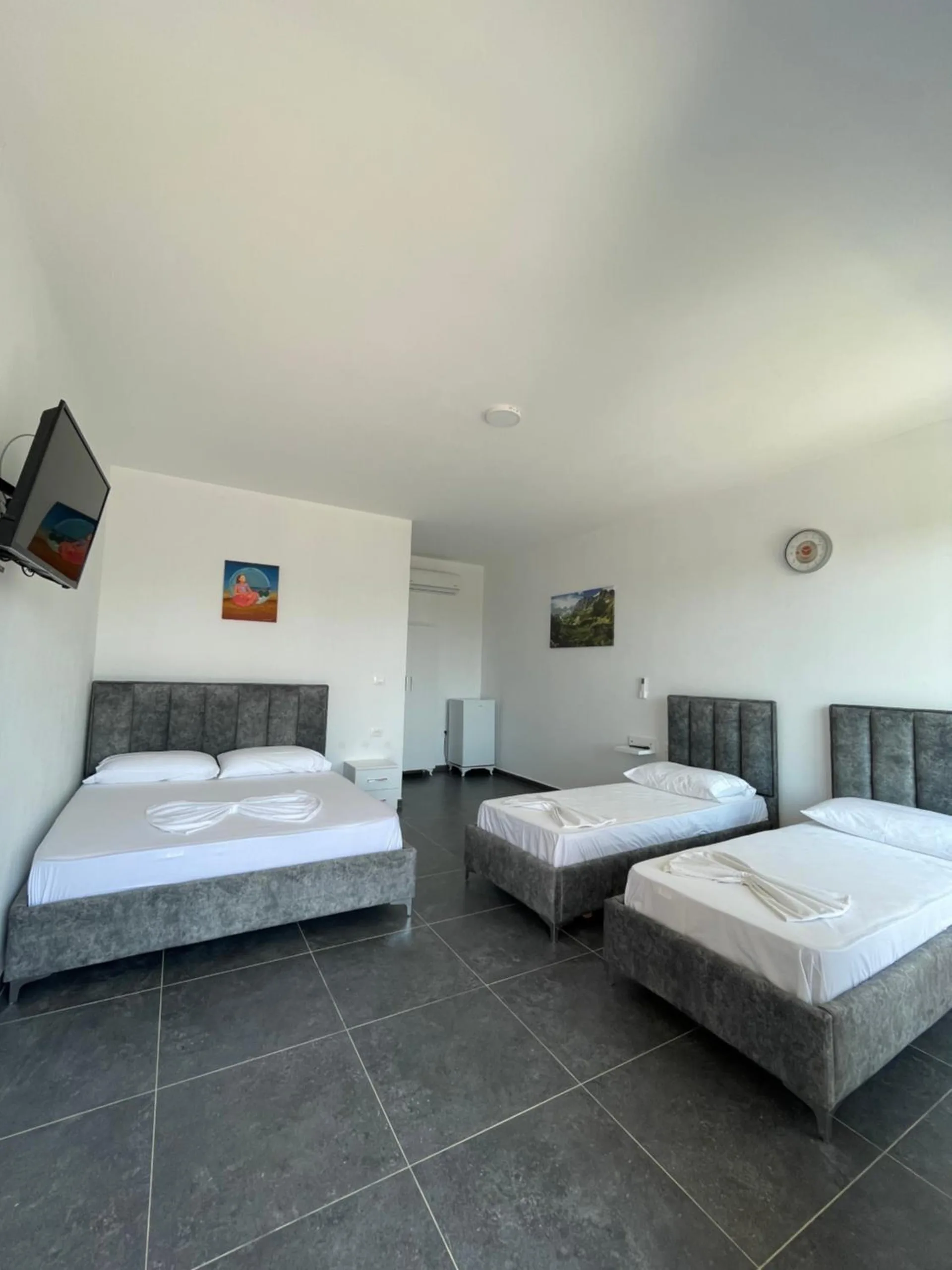 A large white room with a double bed and two single beds in grey velvet frames on a dark stone-effect tiled floor, with a wall television and air conditioning