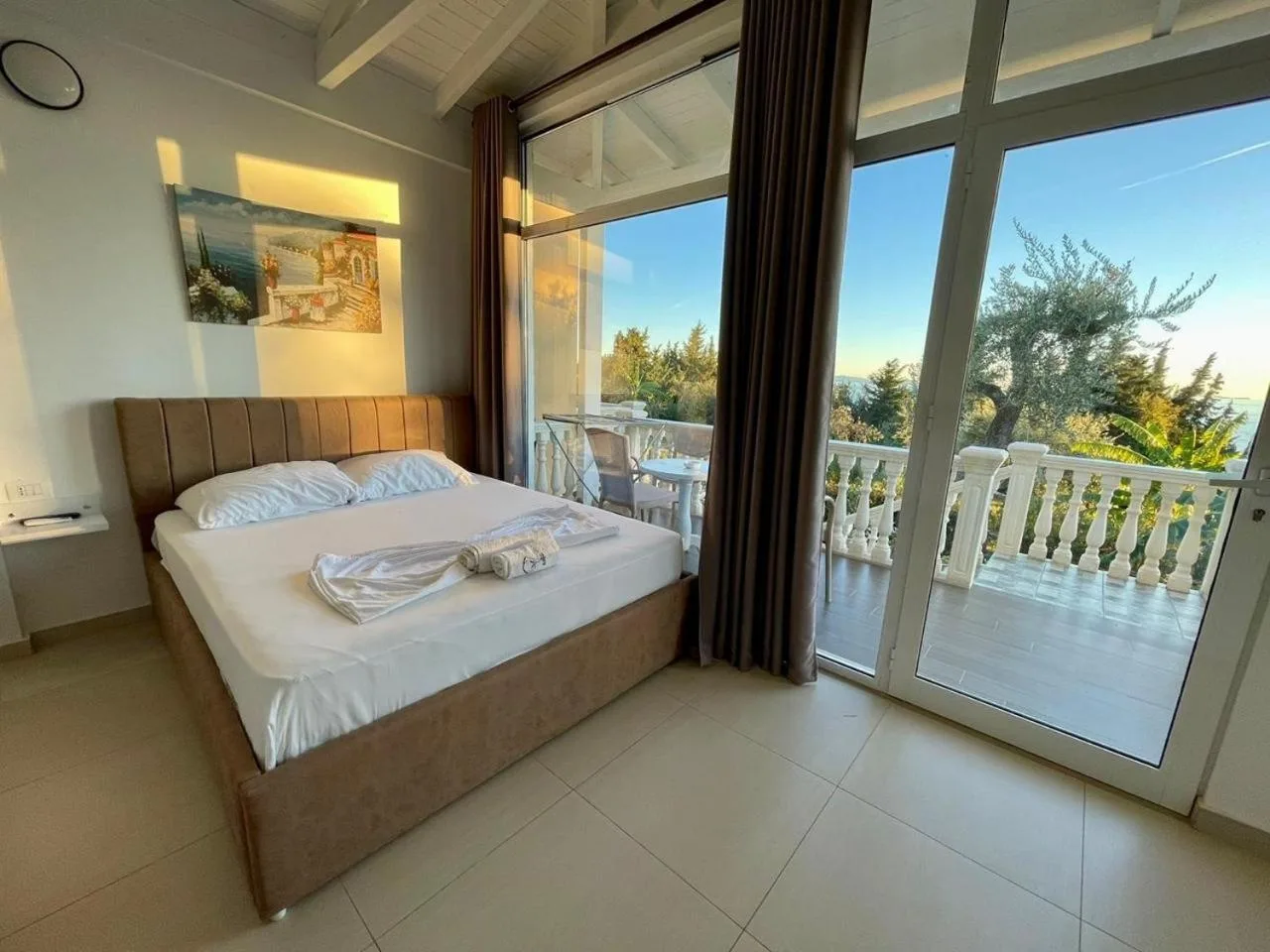 A double bed with a padded headboard in evening light, beside floor-to-ceiling glass doors onto a balustraded porch and the garden