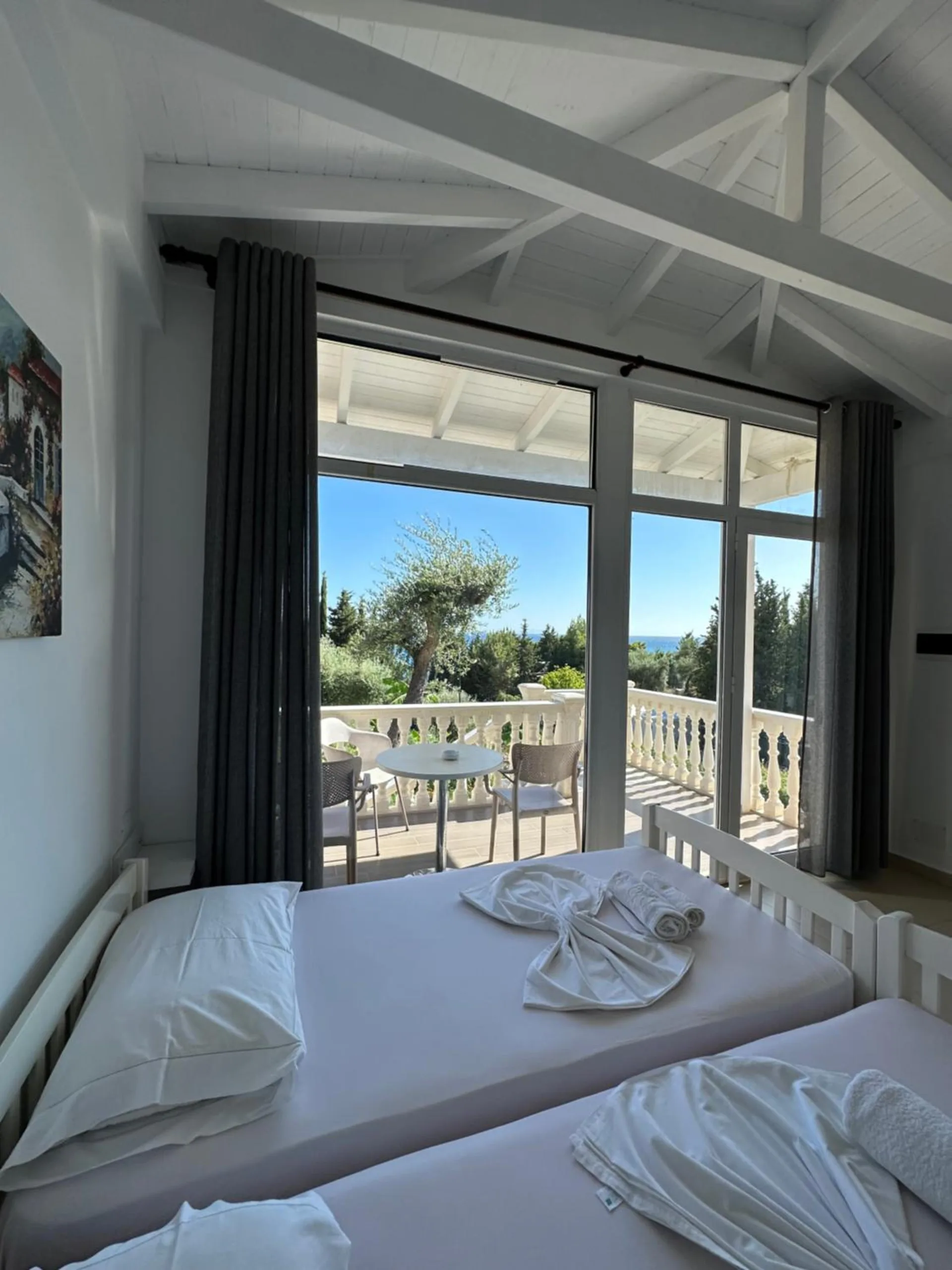 White single beds inside a vaulted room, and through the open glass doors a porch table with chairs, a white balustrade and an olive tree