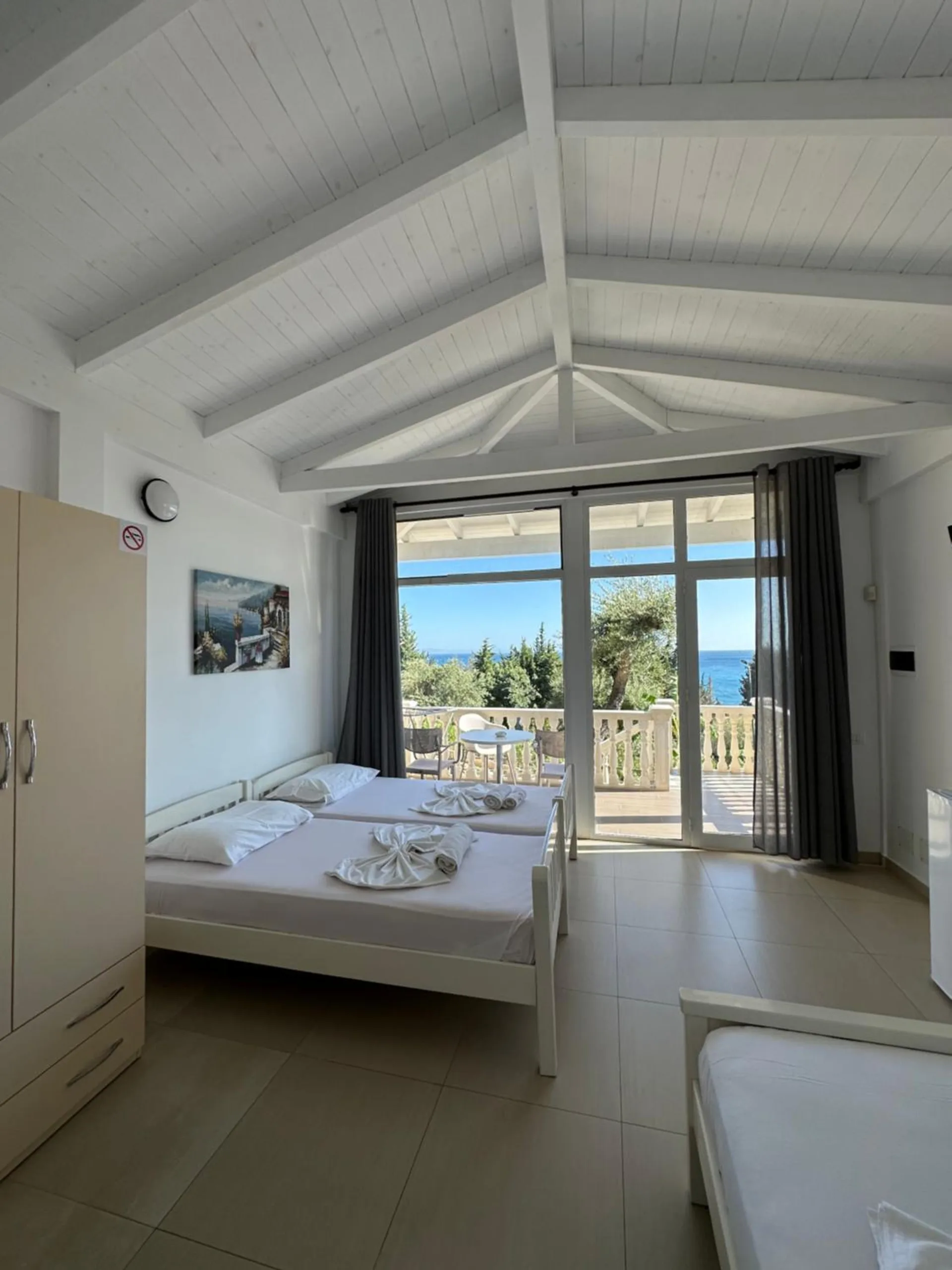 A vaulted white room with beds under a timber truss ceiling and a wide glass door onto a balustraded porch with the sea beyond the olive trees