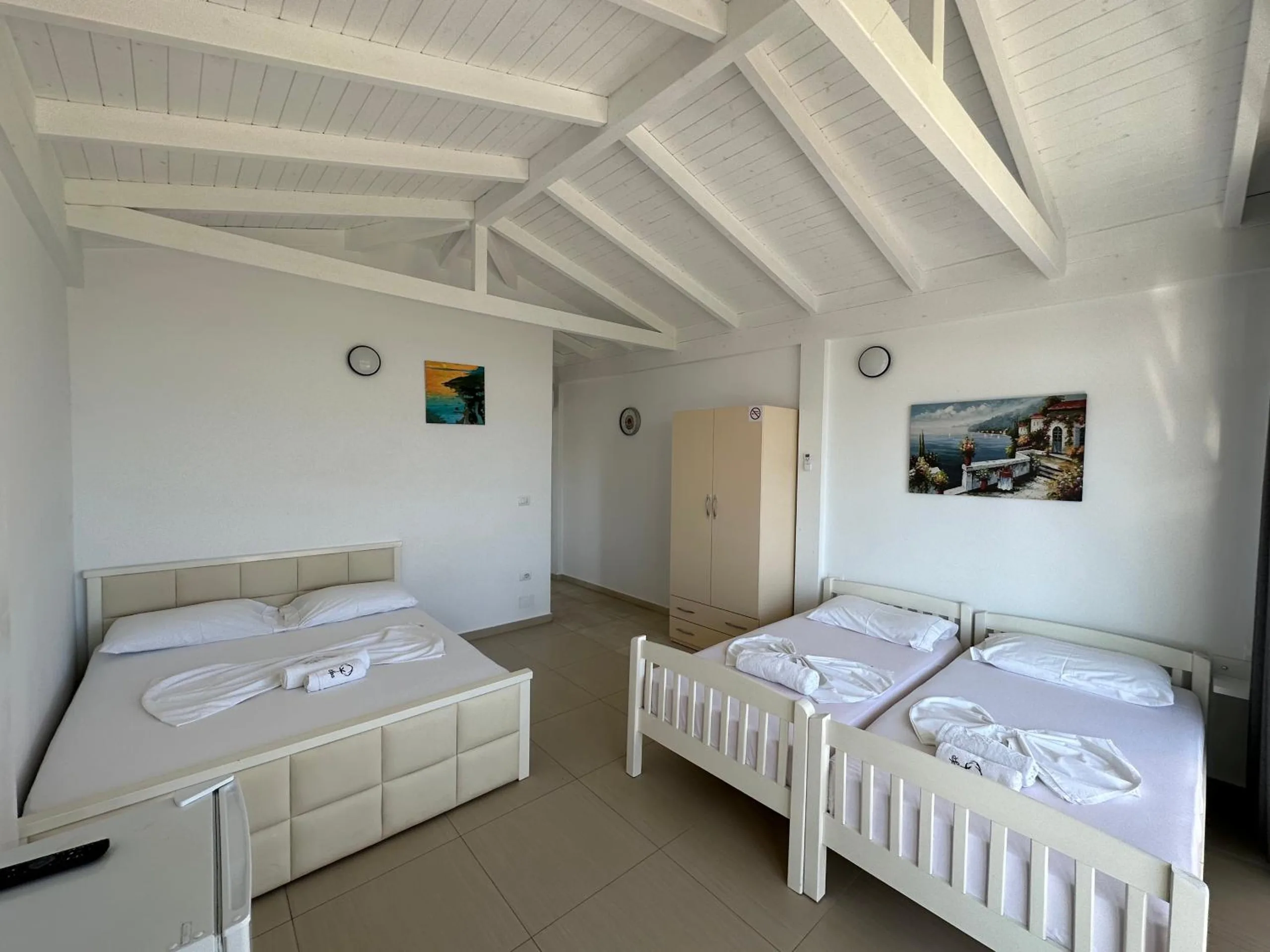 A vaulted room under white-painted timber trusses, with a cream double bed and two white wooden single beds side by side and a pale wardrobe