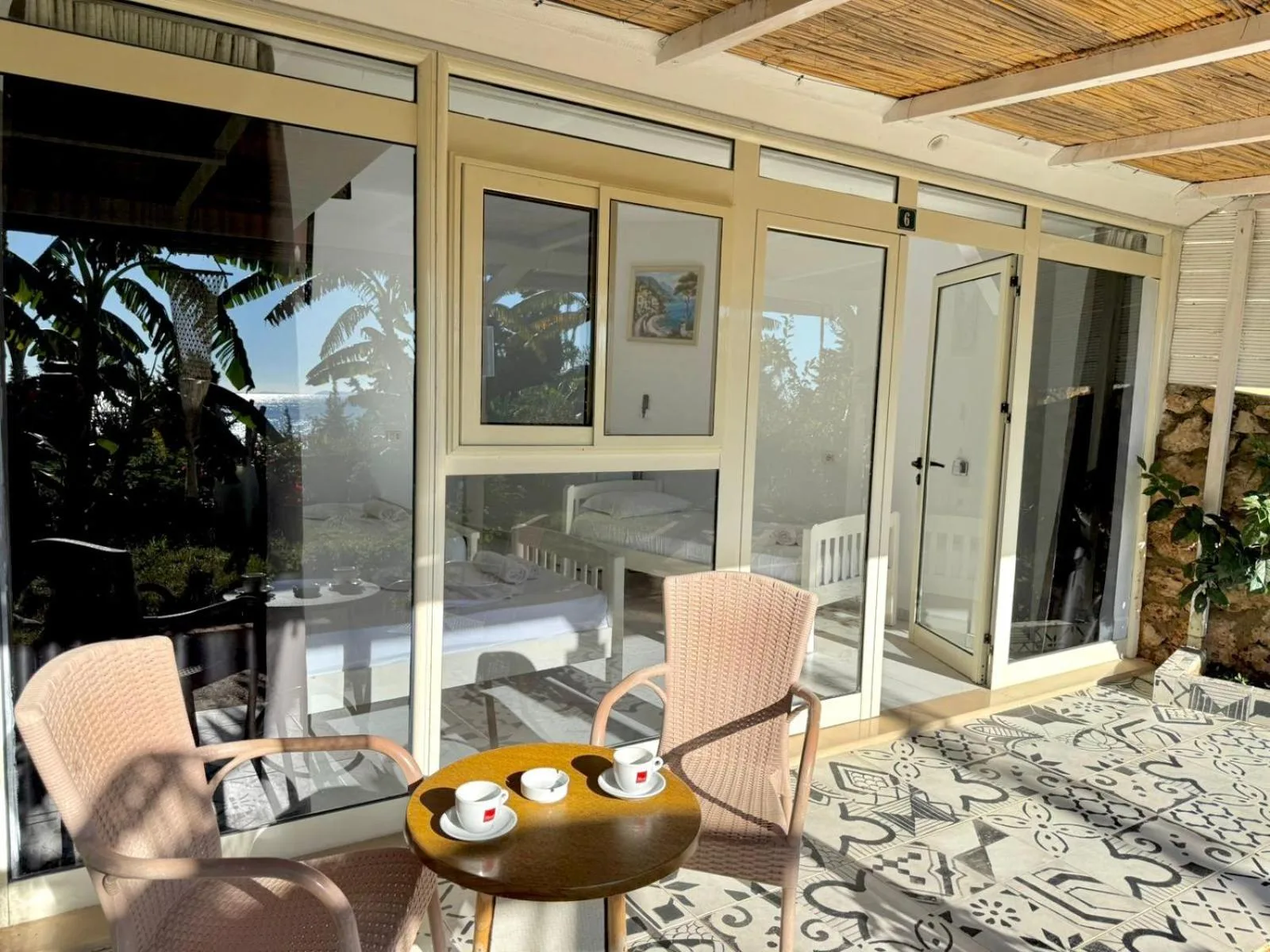 Three cups of coffee on a round wooden table between two wicker chairs on a patterned tile floor, in a glazed veranda with a bedroom behind the glass