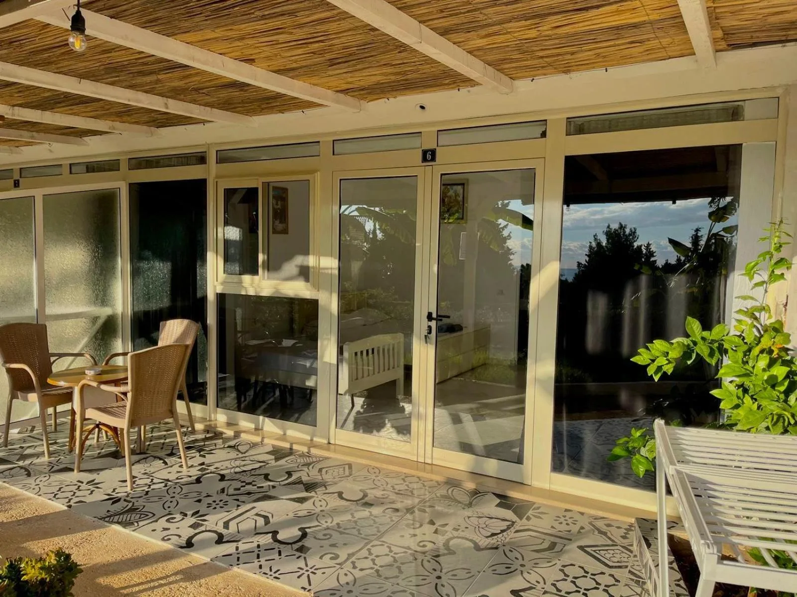 The glass front of a veranda room in low sun, a reed shade overhead, wicker chairs at a table on patterned tiles and a lemon tree in a pot