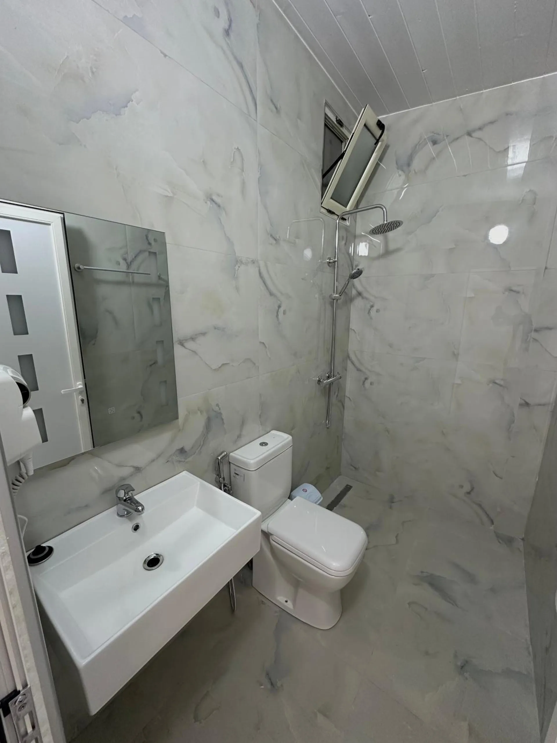 A shower room in white marble-effect tile with a rain shower, a basin, a toilet and a hairdryer by the mirror