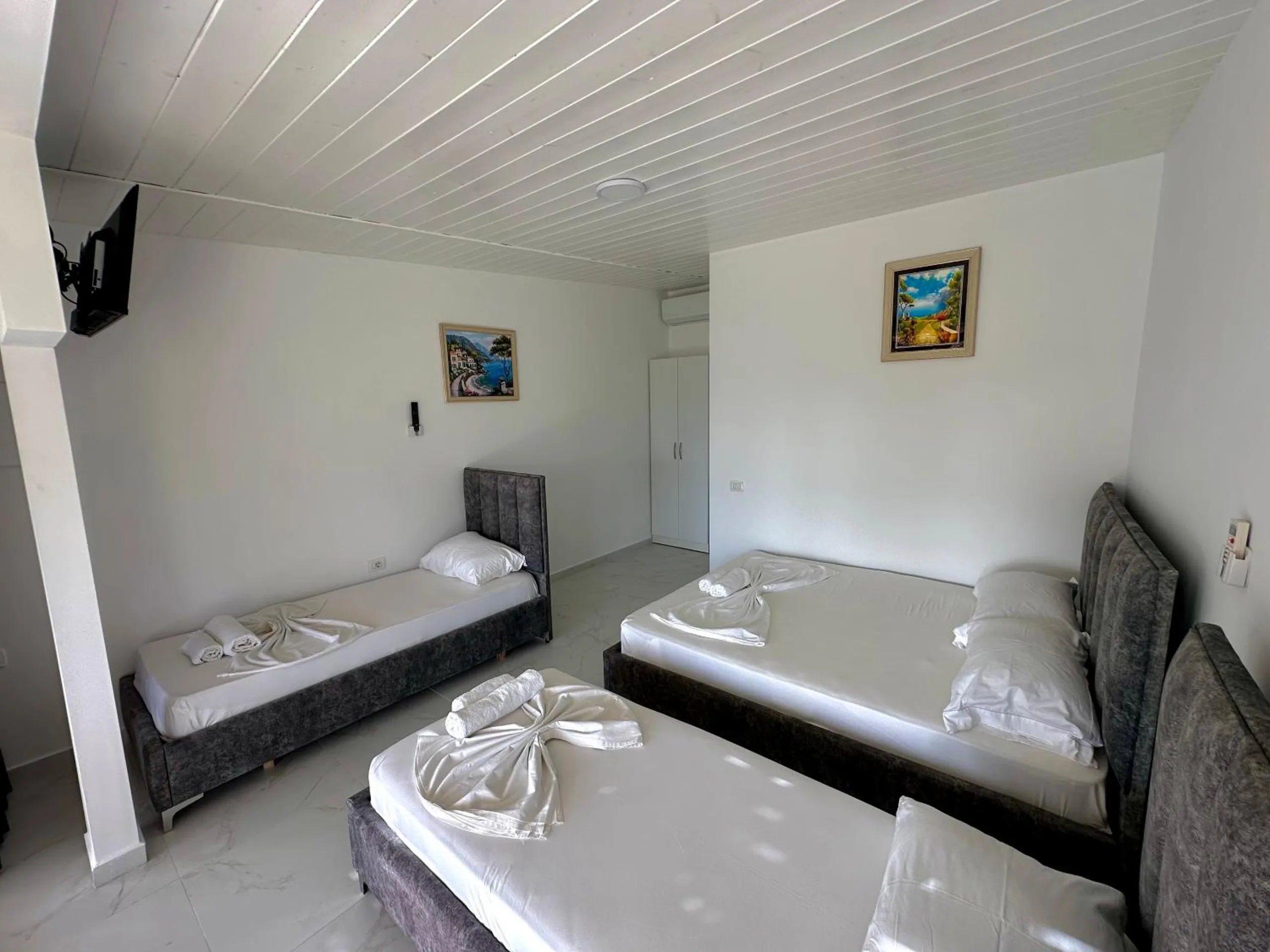 A double bed and two single beds with grey velvet headboards under a white boarded ceiling, a wall-mounted television and framed paintings