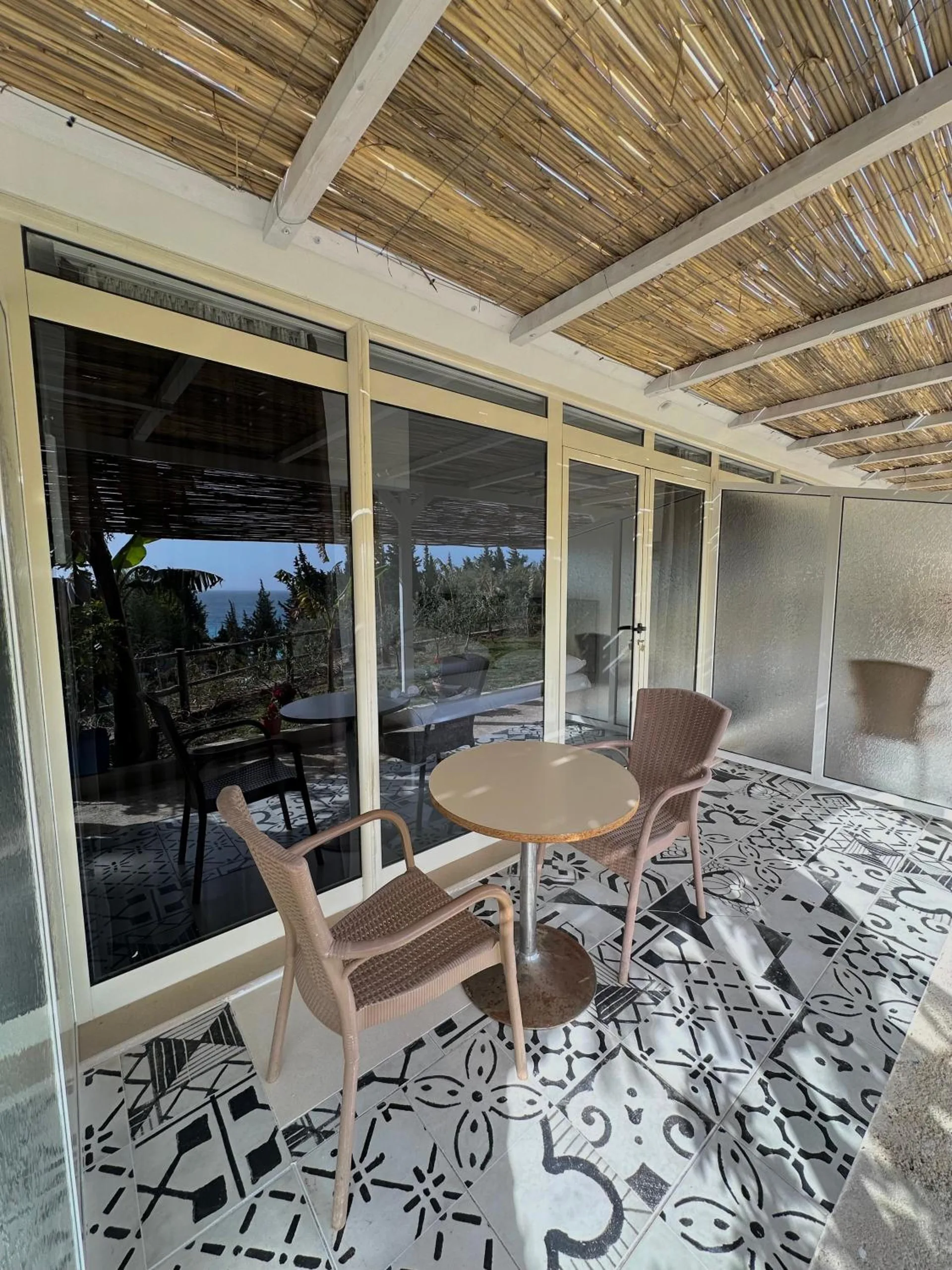 A glazed veranda under a reed-mat shade, floored in black-and-white patterned cement tile, with a round table and two wicker chairs and the sea through the glass