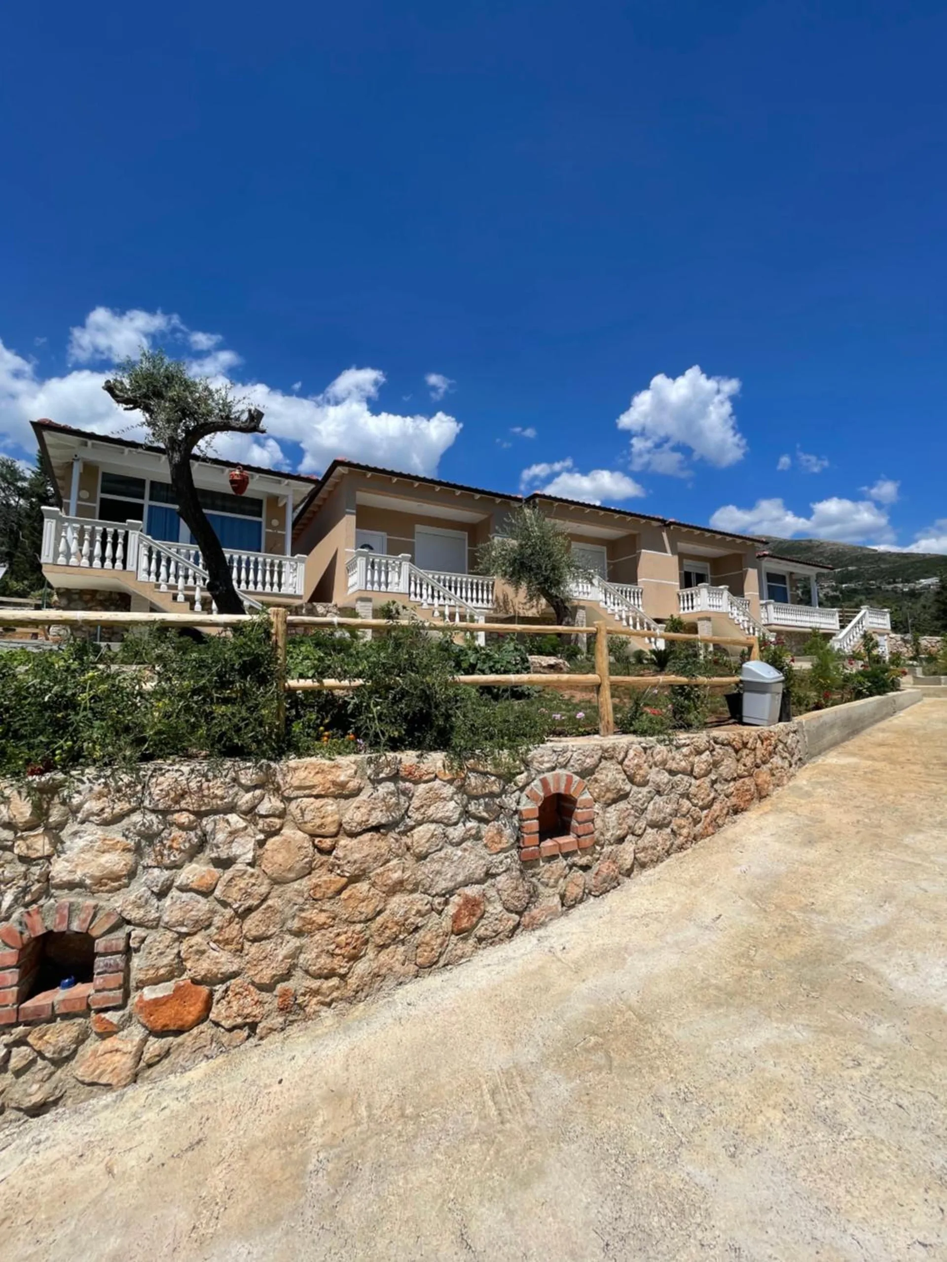 A row of single-storey biscuit-rendered homes with white balustraded porches above a dry stone wall with brick-arched niches, under a deep blue sky