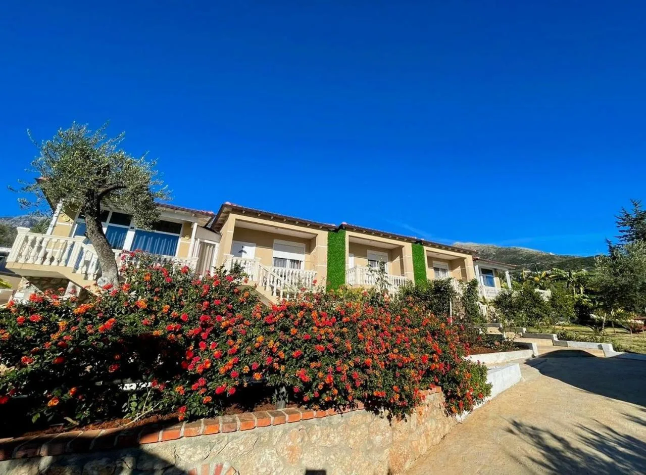 The row of homes with white balustrades behind a bank of red and orange lantana and an olive tree, under a clear blue sky