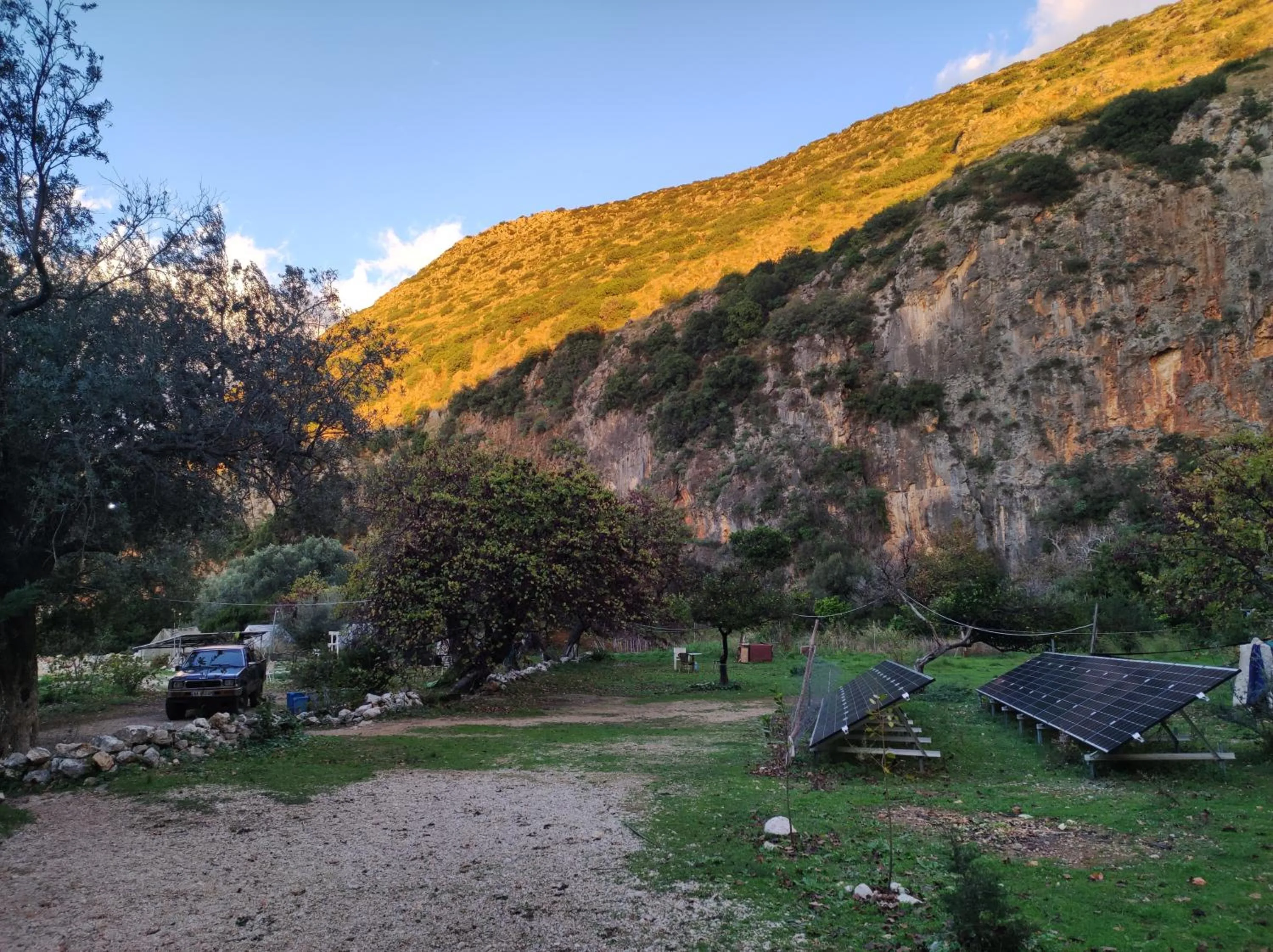 Evening light on the upper wall of Gjipe canyon above the campground meadow, with two rows of solar panels on the grass, a parked four-by-four and olive trees along the track
