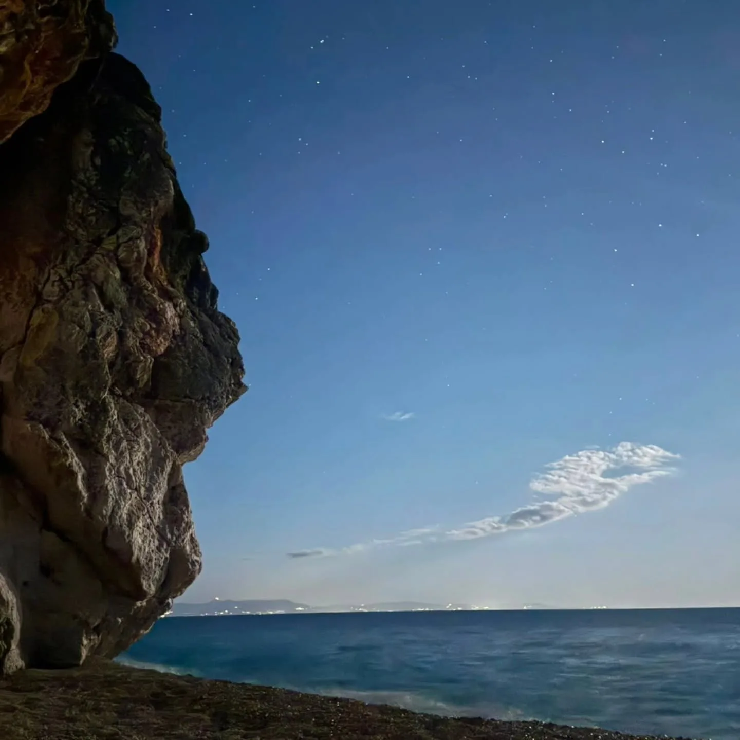Night at the edge of the beach: an overhanging rock face on the left, a few stars in a deep blue sky and the sea flat under it