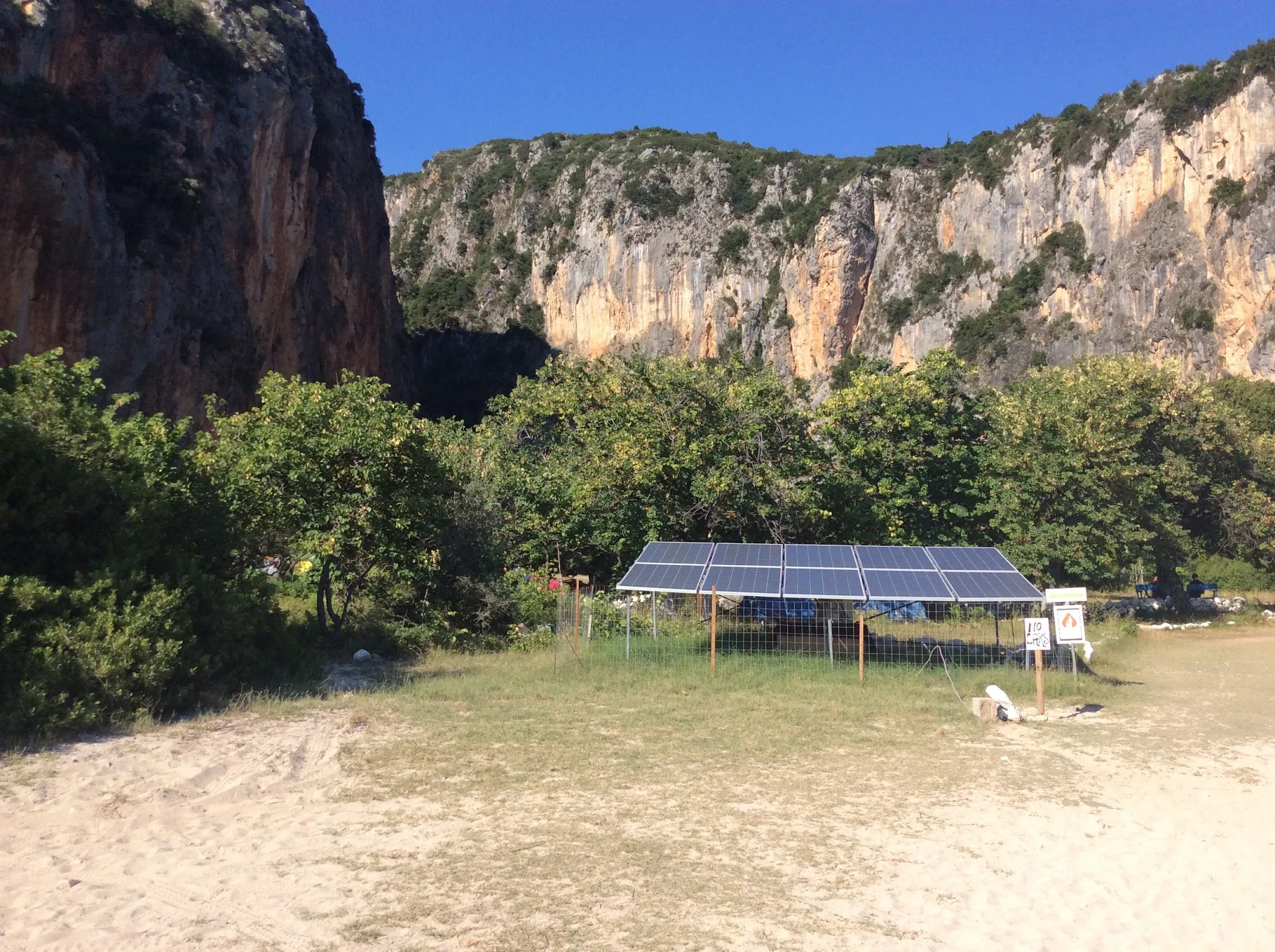 A bank of solar panels on a frame in a fenced meadow at the foot of the canyon, a line of trees behind it and the long pale cliffs of the gorge above