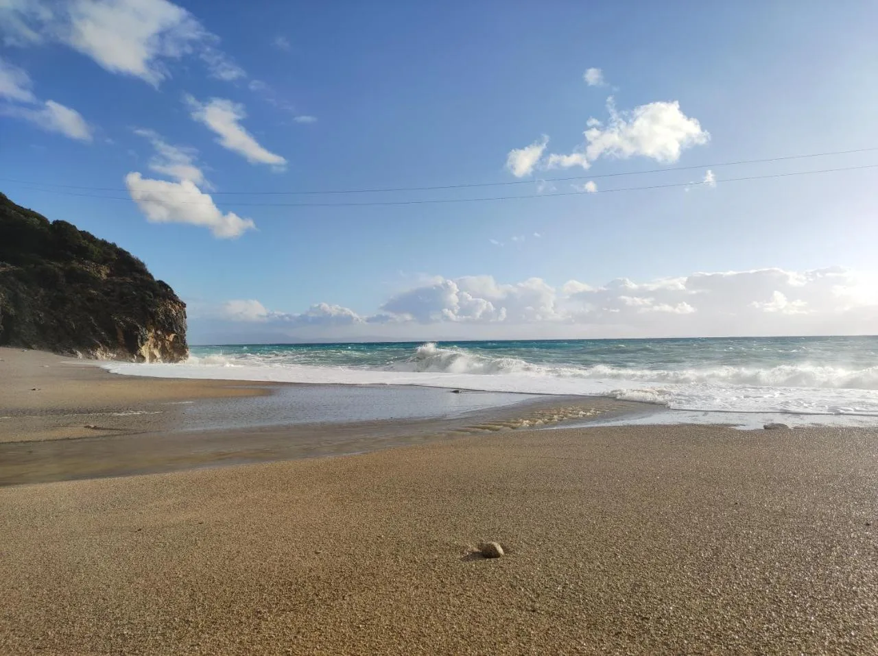 Gjipe Beach on a breezy day, wet sand in the foreground, white surf breaking along the shore and a dark headland at the far end under a blue sky