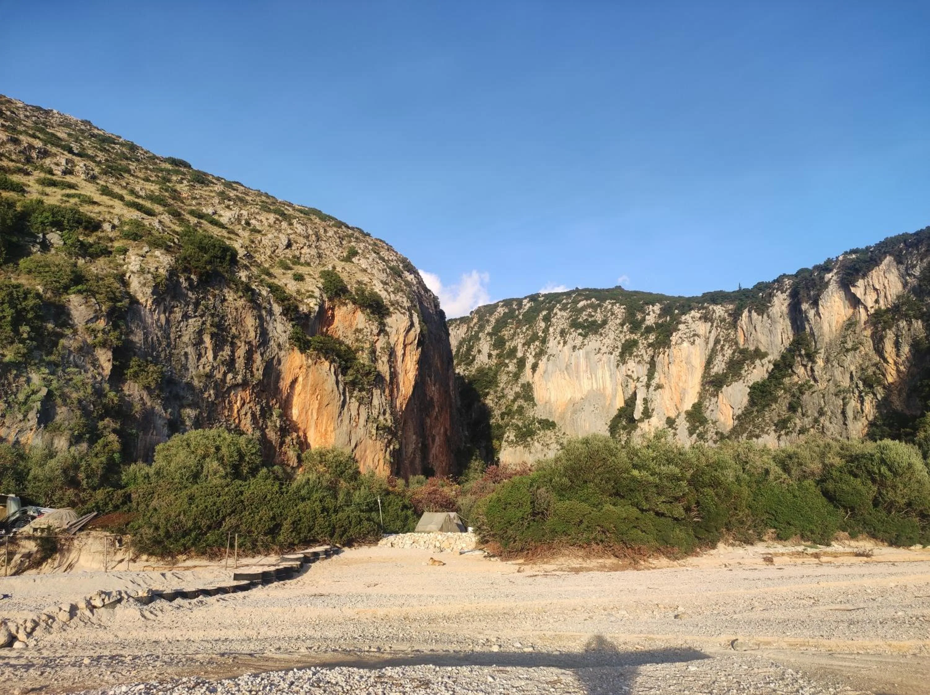The mouth of Gjipe canyon seen from the beach: two sheer orange-streaked limestone walls with a gap between them, scrub and trees at their foot and sand in front