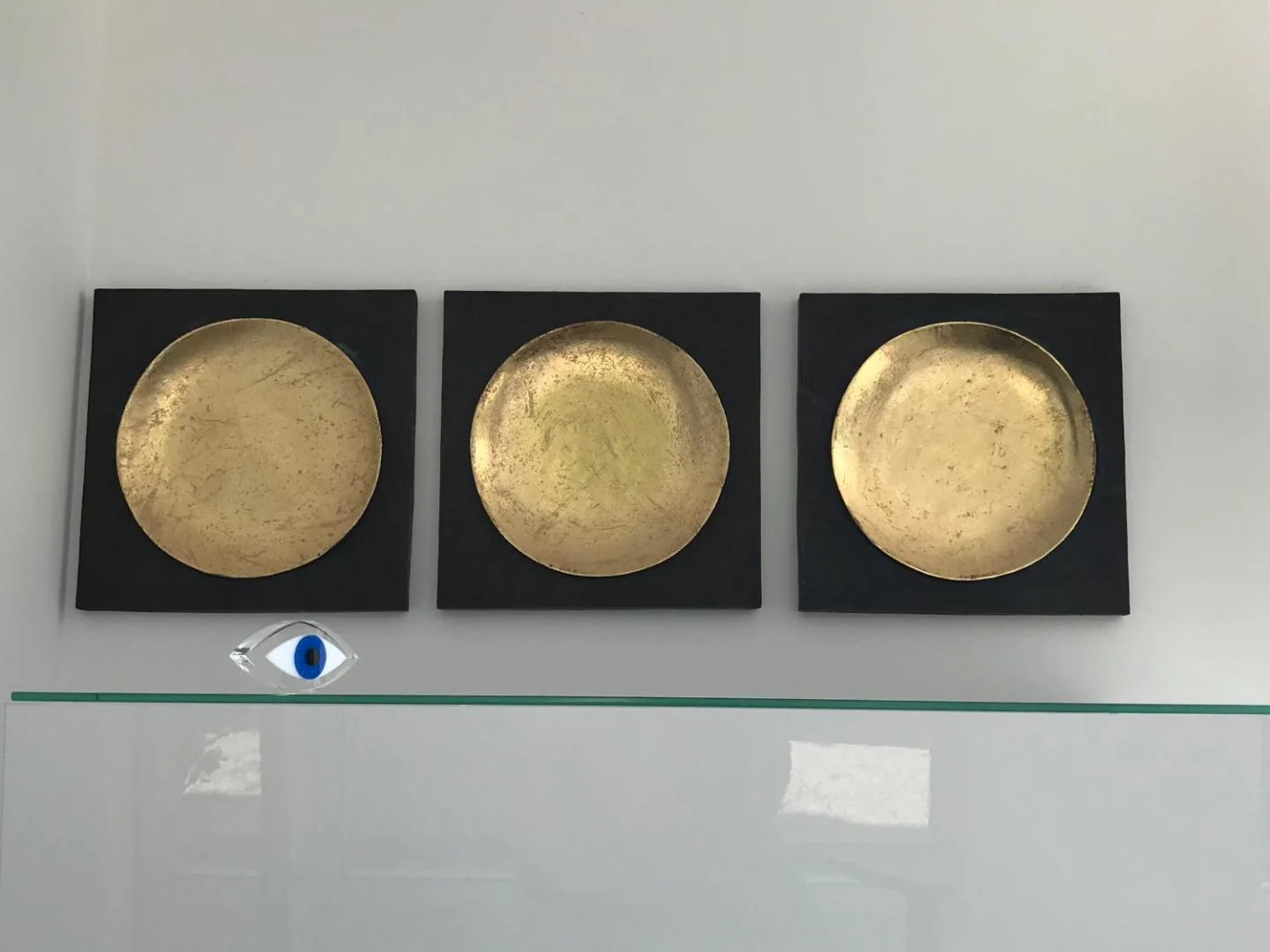 Three hammered brass discs mounted on black panels on a white wall, a small blue eye charm on the glass shelf beneath