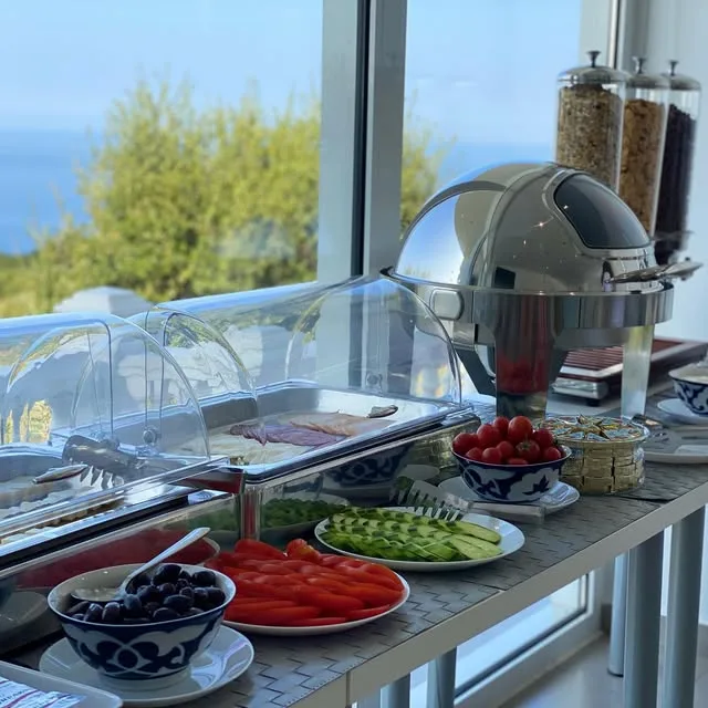 The breakfast table by the window: cereals, a covered hot dish, tomatoes, cucumber and olives, the hillside outside