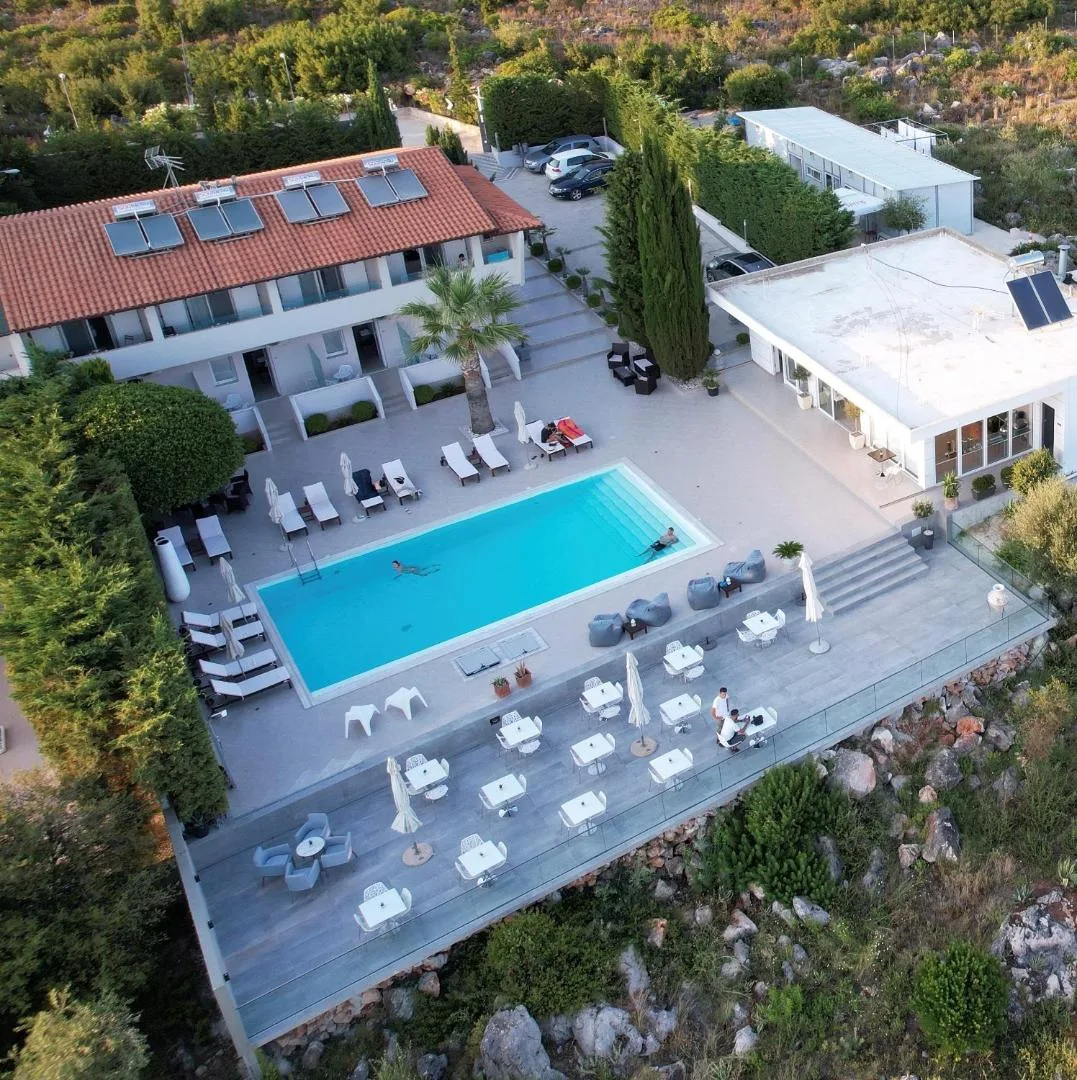 The whole property from the air: the red-roofed block, the pool, a palm, the white terrace laid with tables on the edge of the slope