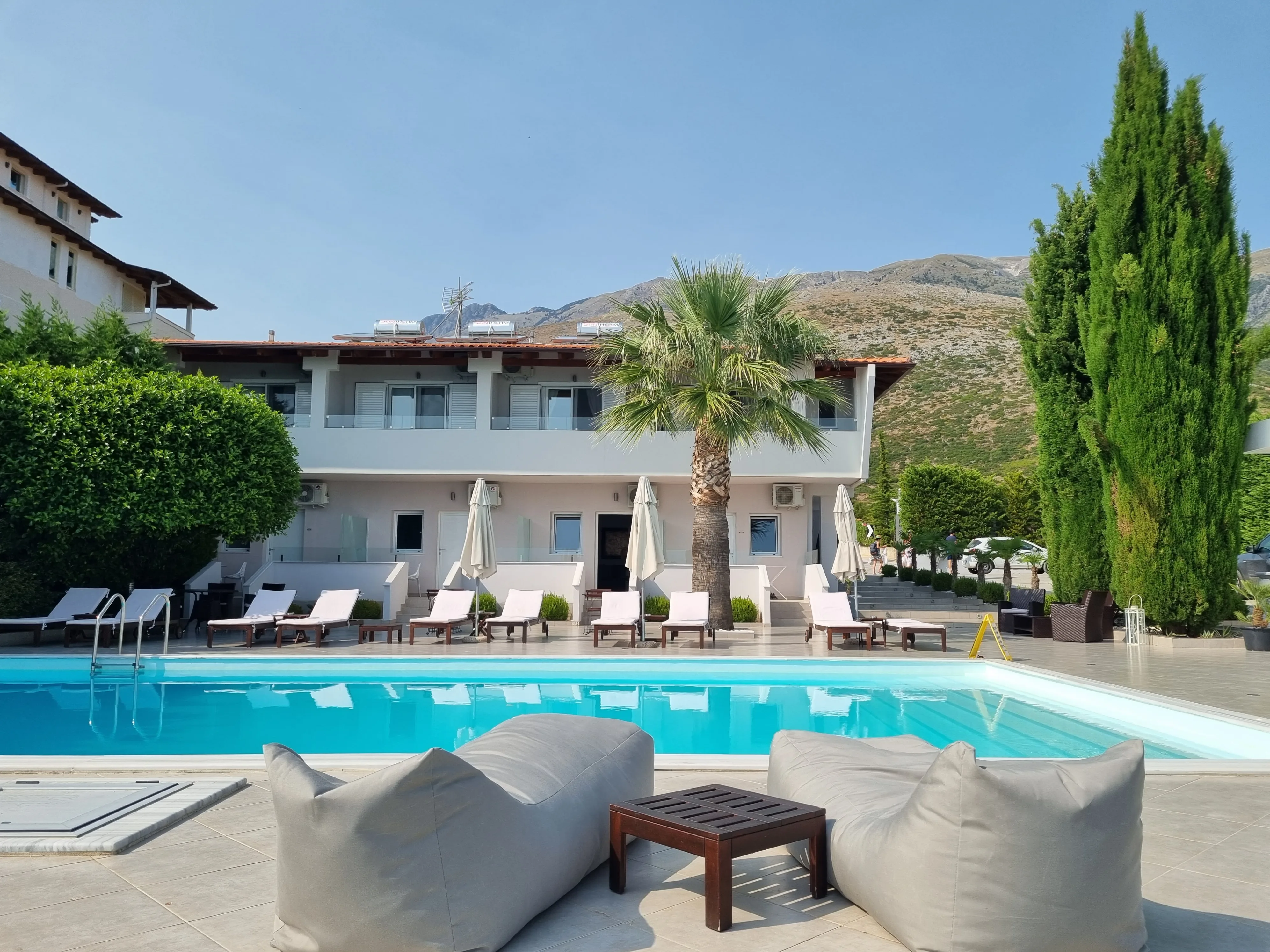 The pool in full sun in front of the two-storey white block with its red tile roof, a fan palm and a tall cypress, the mountain behind