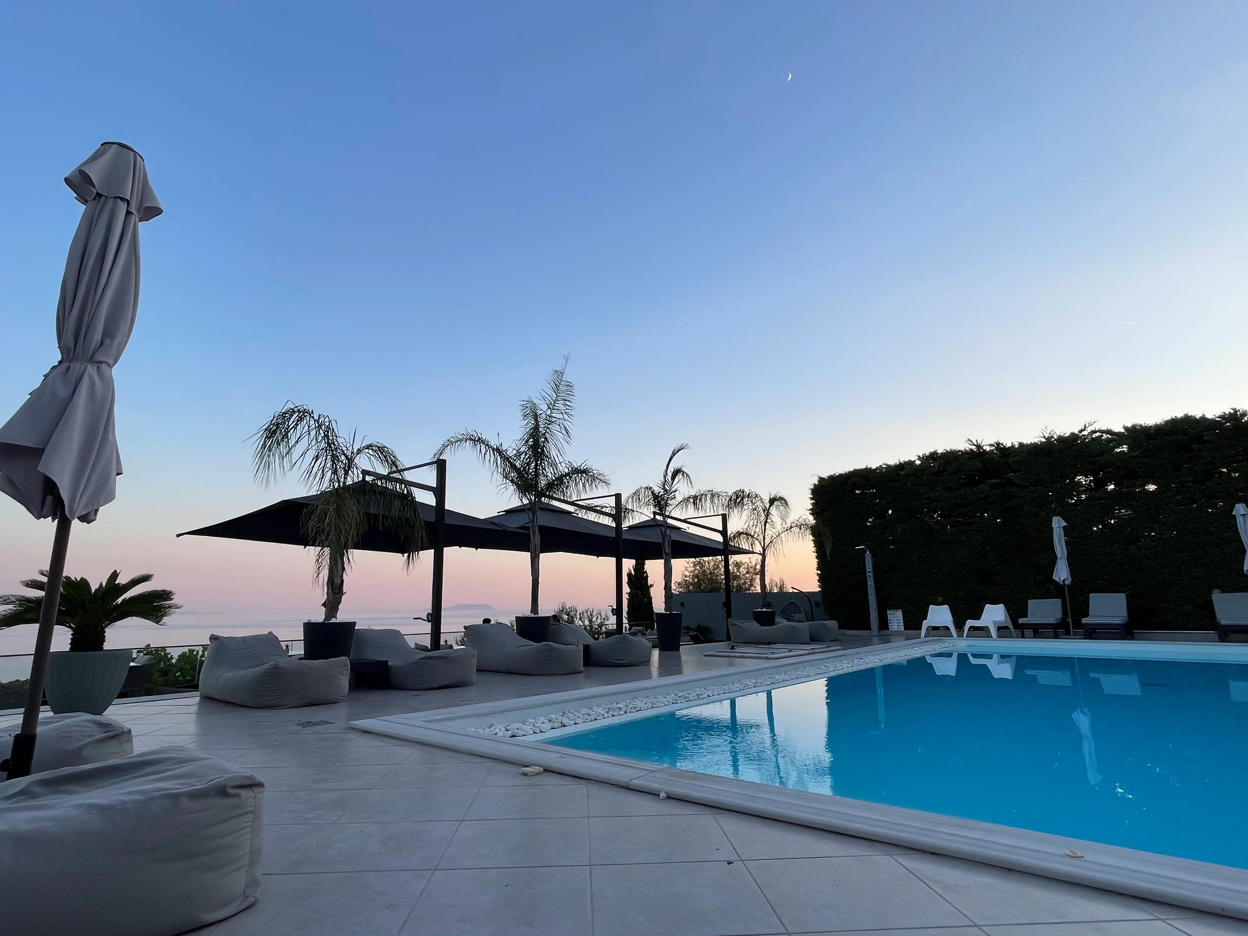 The pool terrace at dusk: a crescent moon in a pale evening sky, a pink afterglow along the sea, an island low on the horizon, fan palms and black sun canopies over the loungers