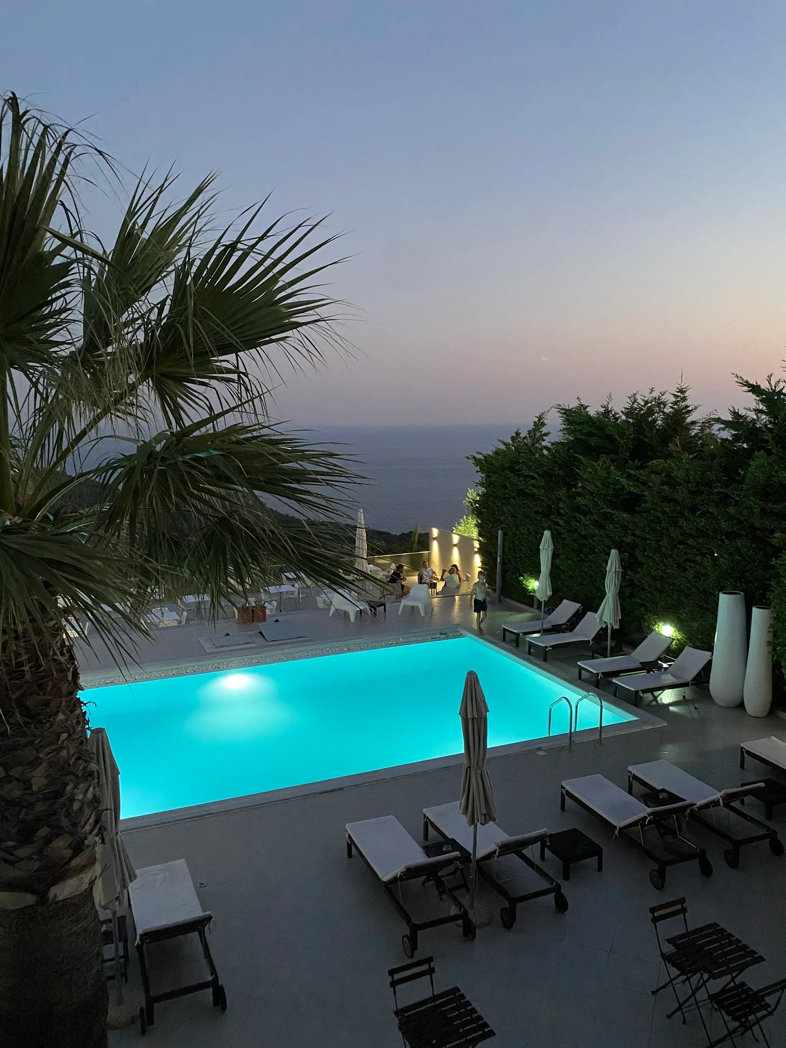 The pool lit turquoise at dusk seen from an upper floor, palm fronds in the foreground and the sea dark beyond the hedge