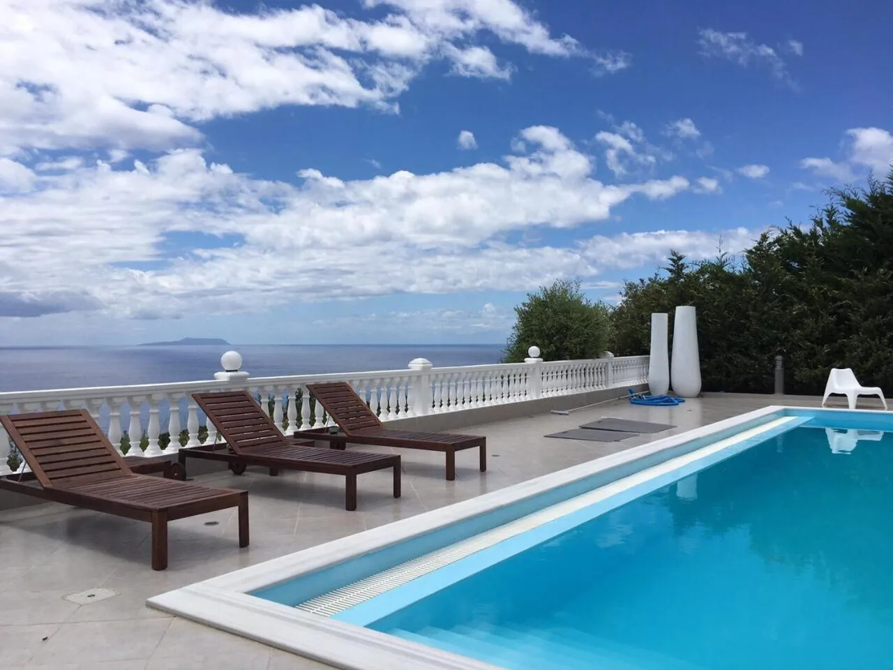 Three wooden loungers on the pool deck beside a white balustrade, the sea and an island beyond under summer clouds