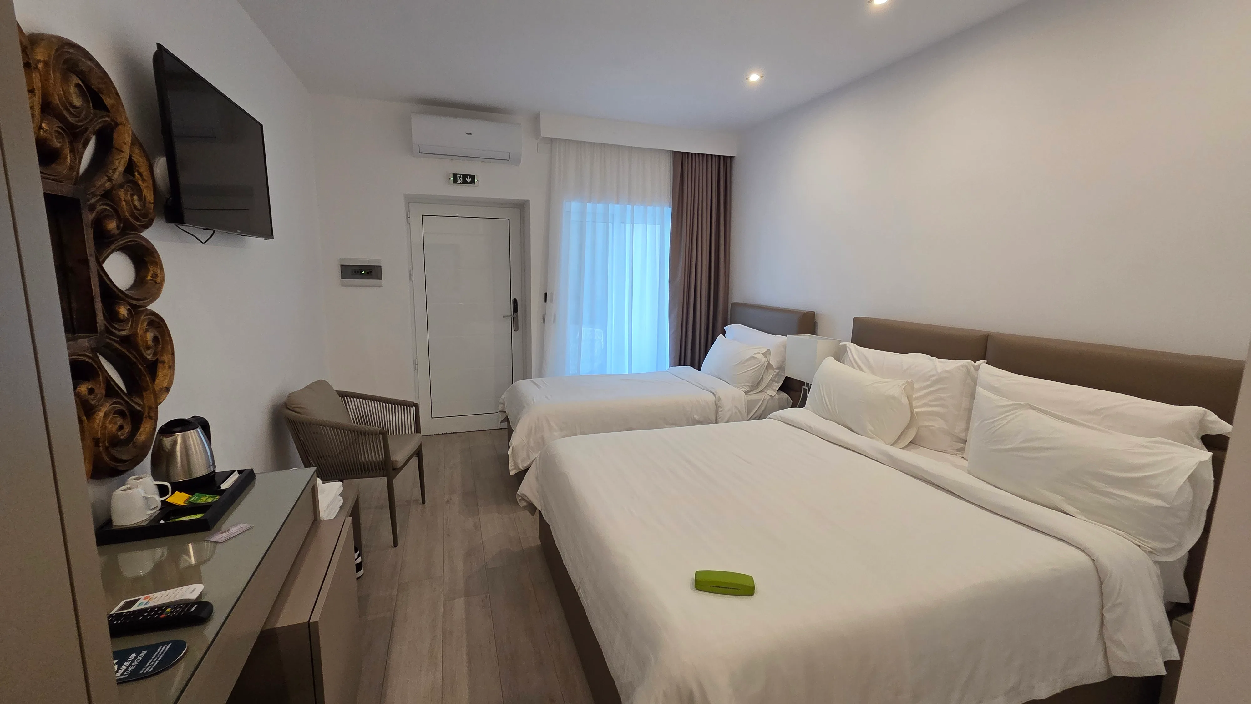 A bright room with a double and a single bed, a kettle and cups on the desk and a carved wooden panel on the wall