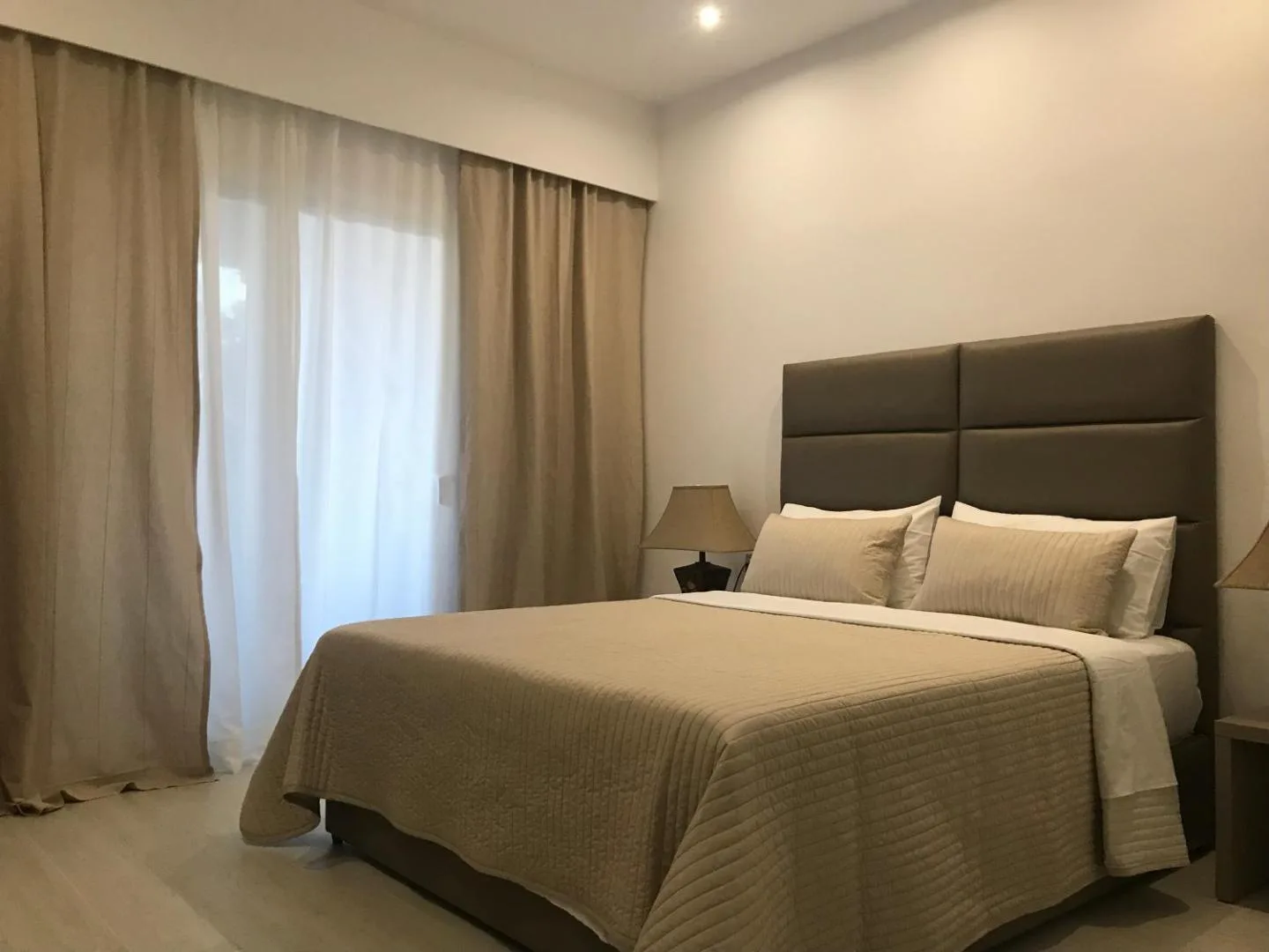 A double room: a tall padded headboard, sand-coloured bedspread and long curtains over the balcony door