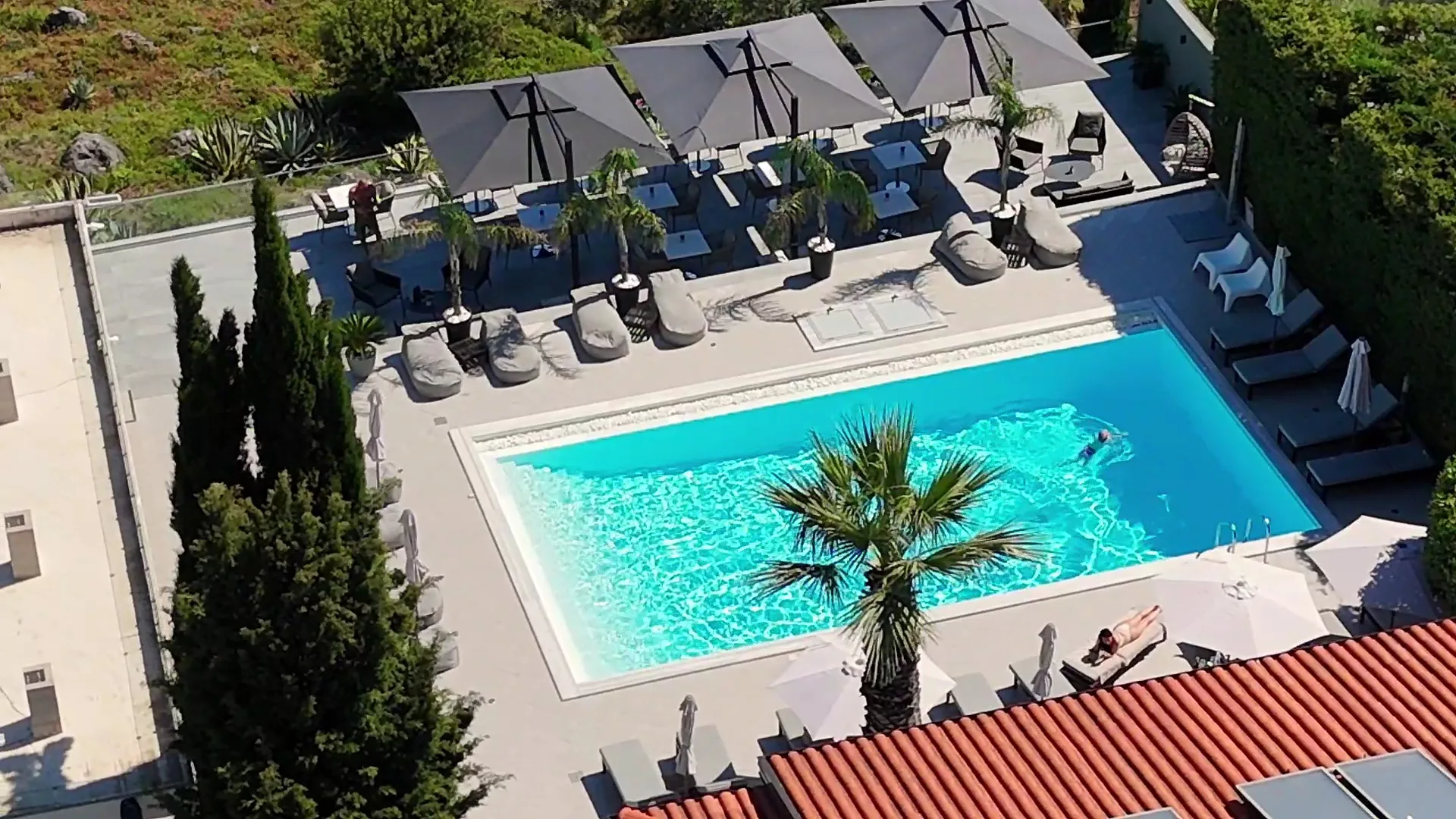 The pool from the air, a palm on its island in the middle, loungers and grey sun canopies around it and the red roof in the corner