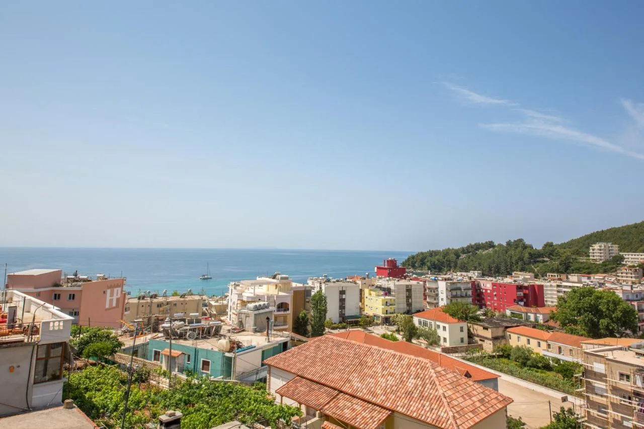 The view from an upper floor over terracotta roofs and Himarë's blocks to the open sea and a sailing boat