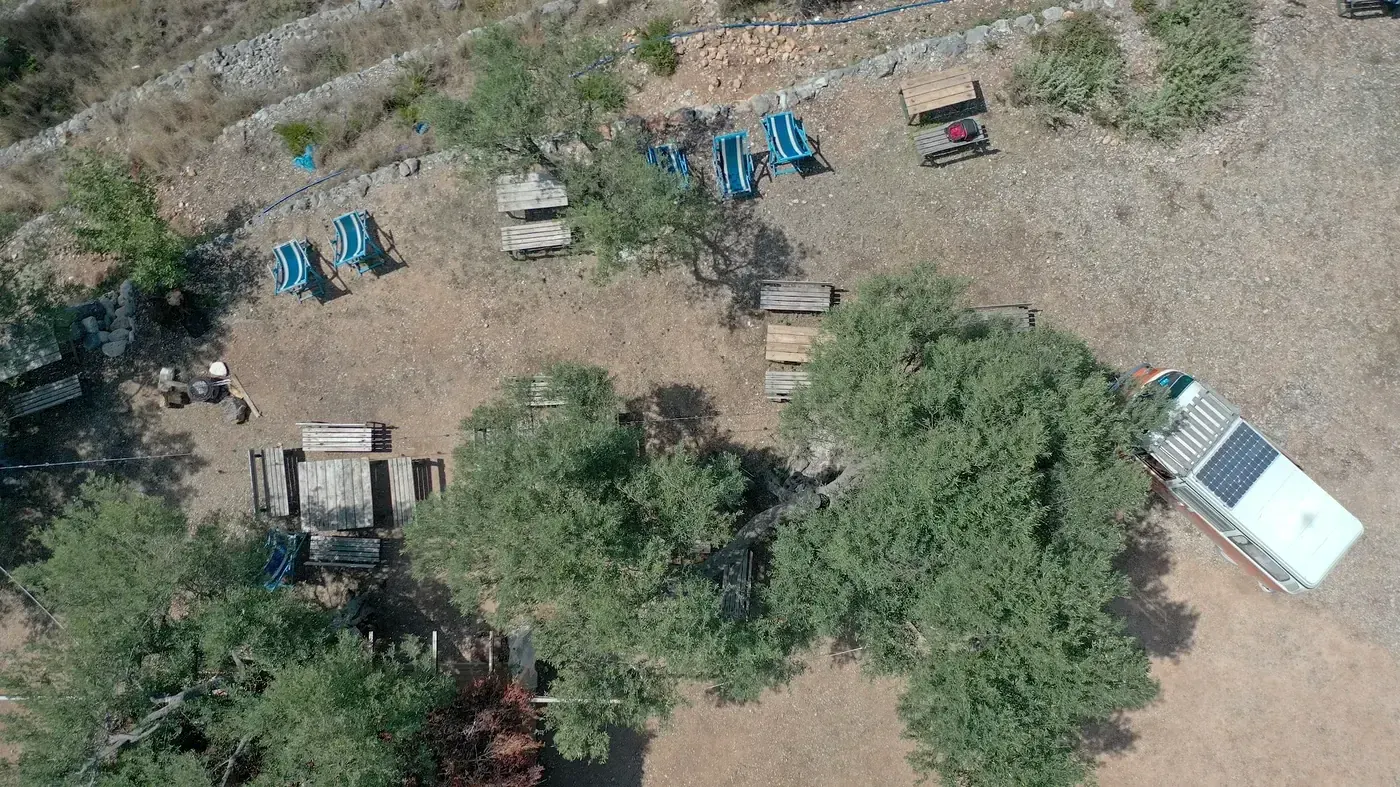 Looking straight down on a pitch at Camping Himara: deck chairs, pallet tables, an old olive and a white campervan on the gravel