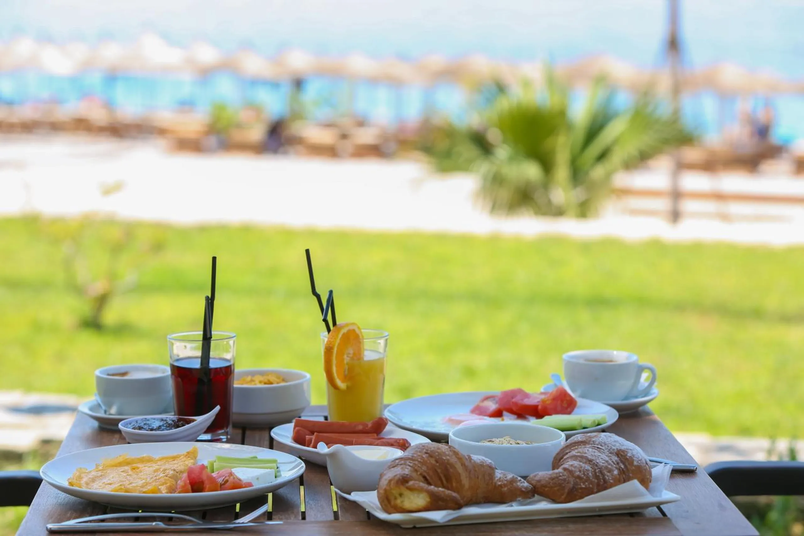 Breakfast laid on a table at the edge of the lawn, croissants and orange juice, the umbrellas of the beach out of focus beyond