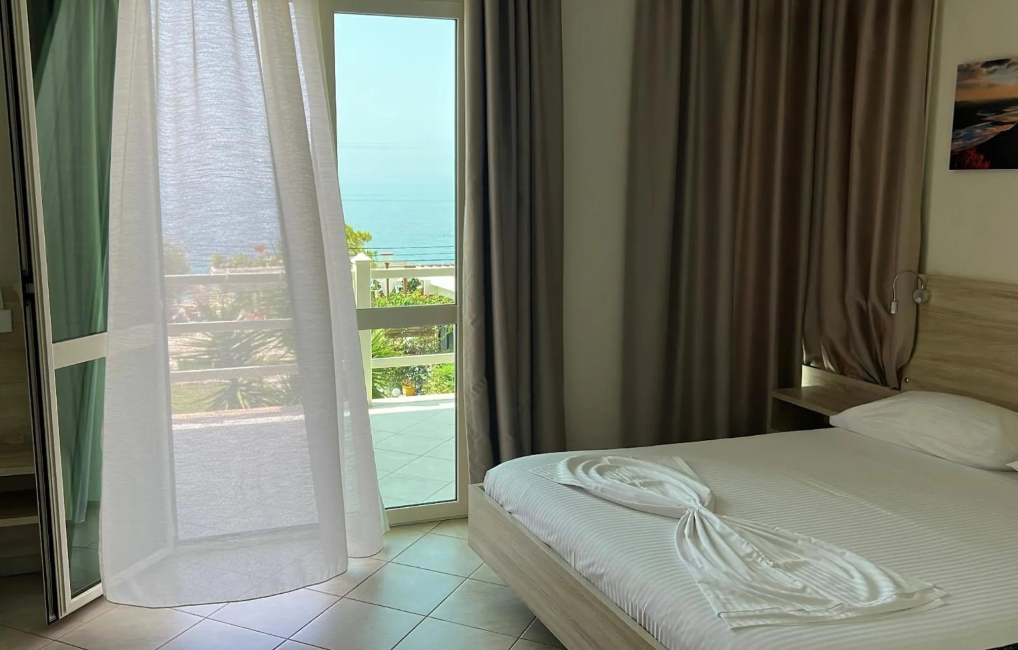 A double bed beside an open balcony door, white curtains moving and the sea beyond the garden