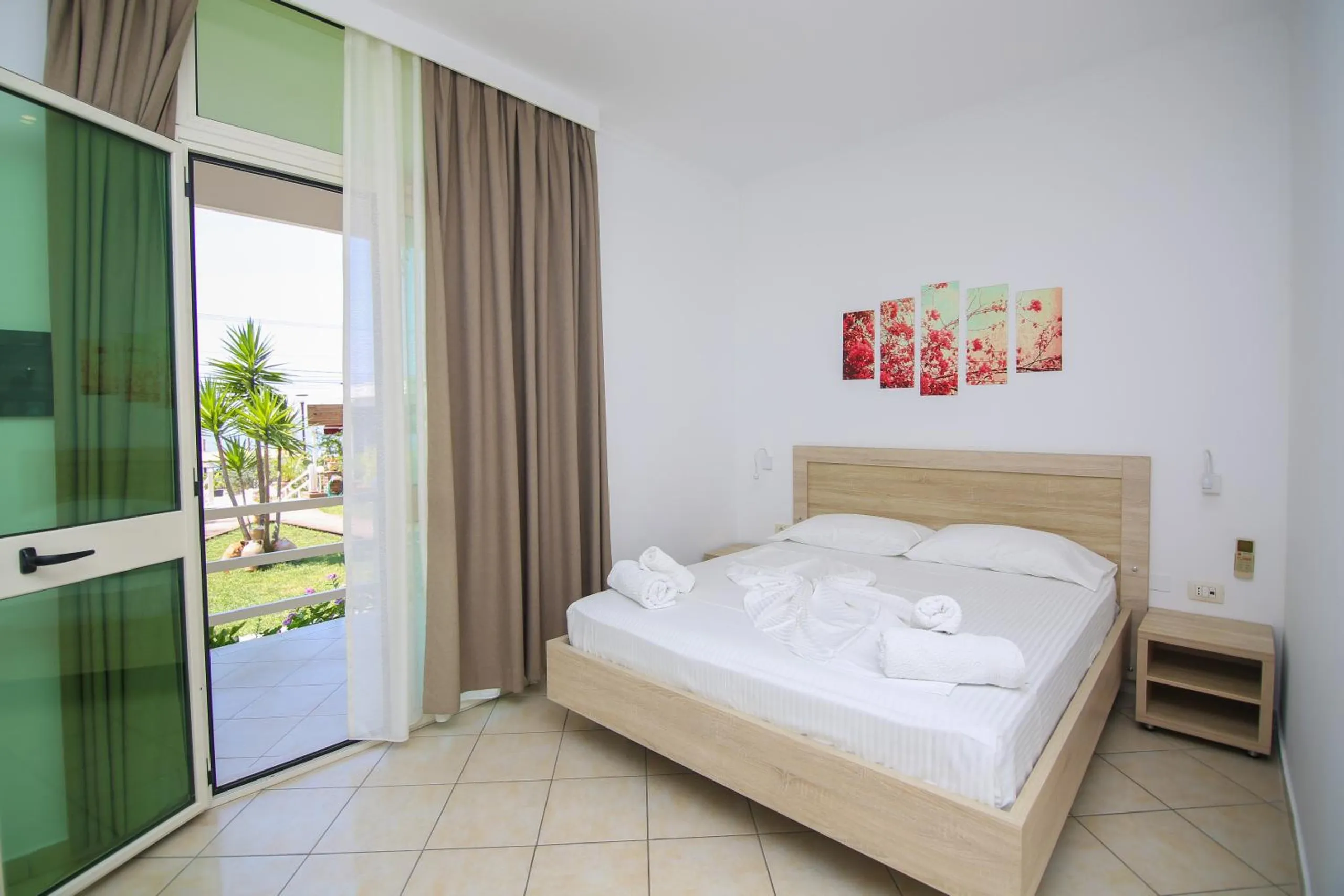 A double room with its green glass door open onto the balcony and the garden, white walls and a pale-oak bed